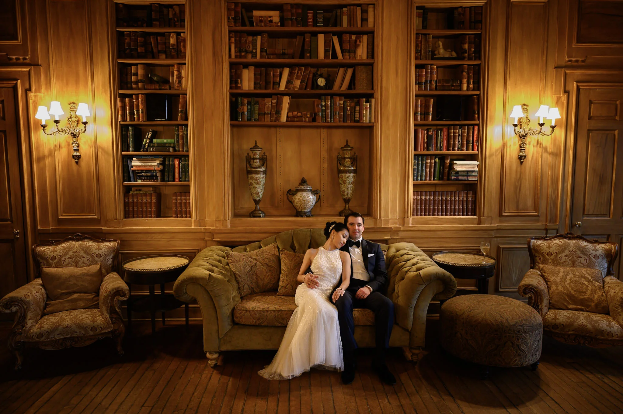 A bride and groom sitting on a vintage green sofa in a wood-paneled room with a large bookshelf and ornate lighting fixtures. The bride is resting her head on the groom's shoulder, and they are dressed in wedding attire.