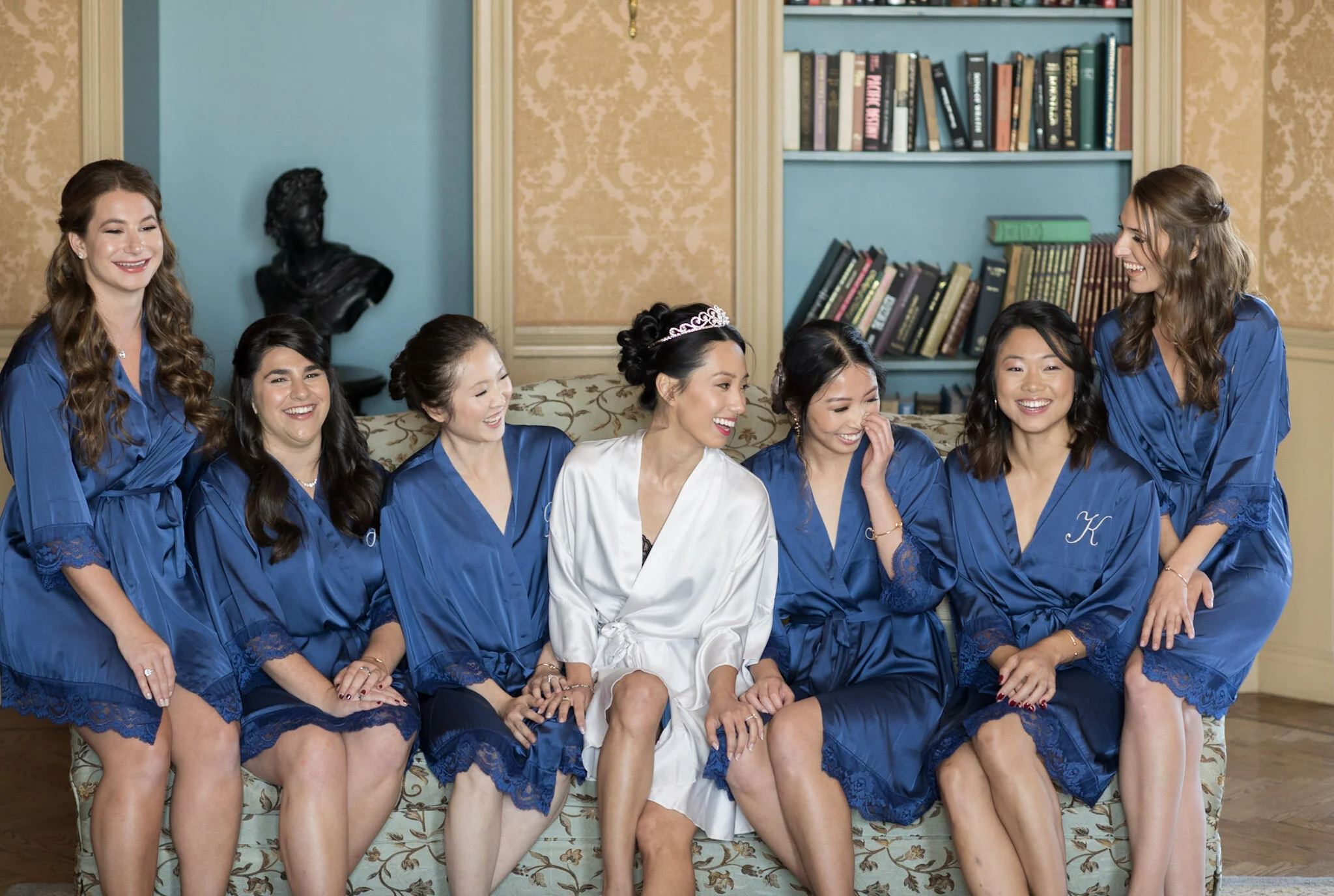 A bride in a white robe wearing a tiara surrounded by eight bridesmaids in matching blue robes, sitting and laughing together on a floral couch in a room with bookshelves and a textured wall.