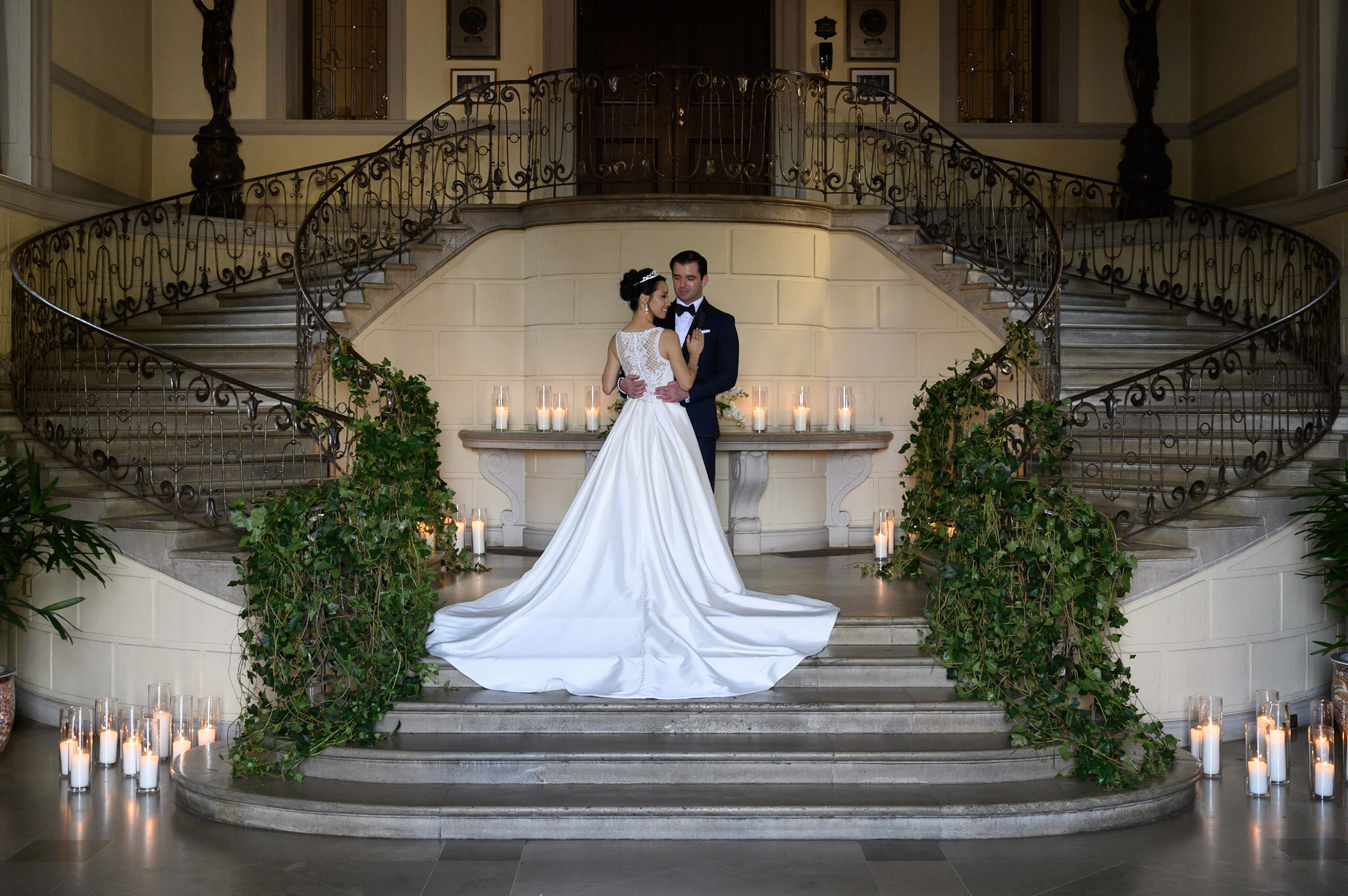 A bride and groom standing on a grand staircase, surrounded by candles and greenery, in an elegant indoor setting with ornate staircase railing and framed pictures on the wall.