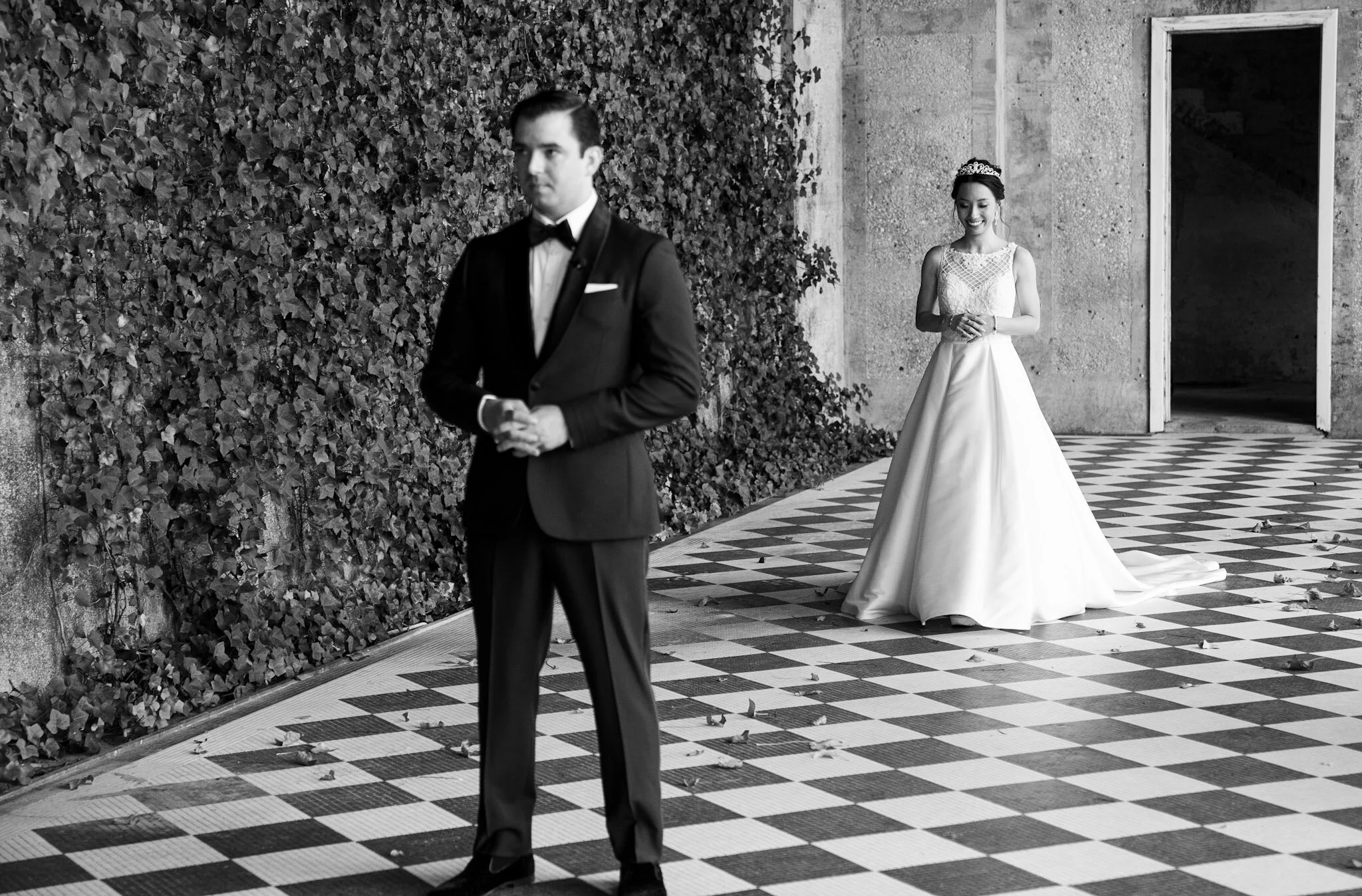 A black-and-white photo of a groom walking away and a smiling bride standing in the background at a wedding. The groom is dressed in a tuxedo with a bow tie, while the bride wears a wedding dress and tiara, standing behind him with her hands clasped.