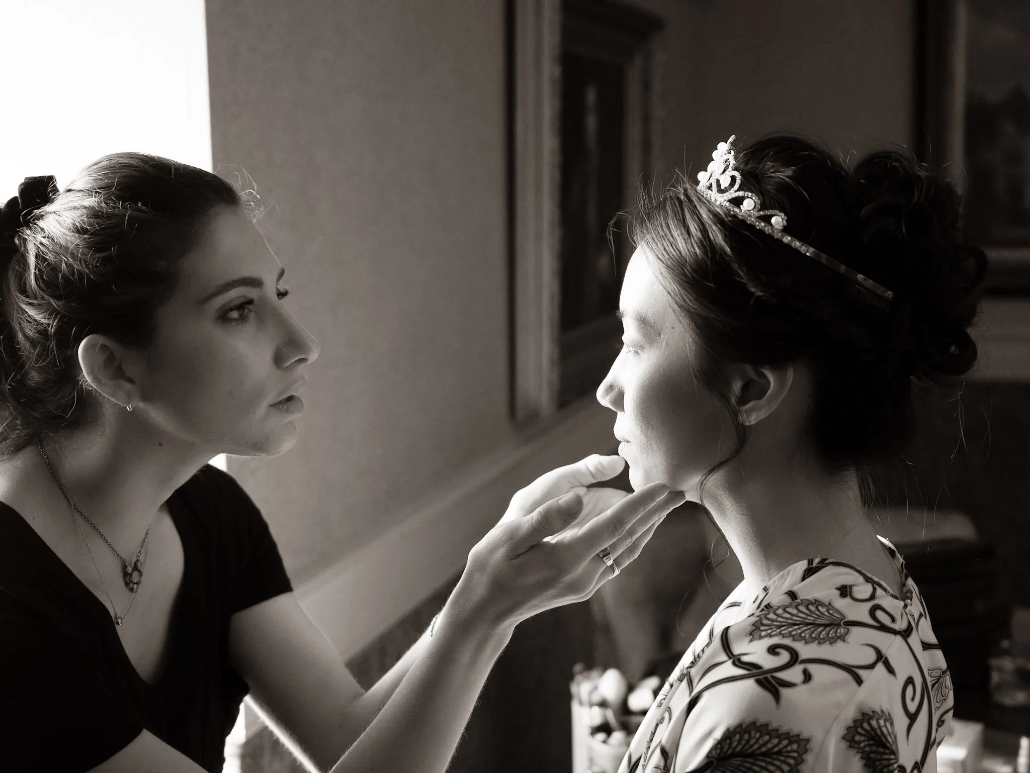 A woman with a hairstyle and a necklace applying makeup or lipstick to a young girl with a decorative headband and a patterned sleeve shirt.
