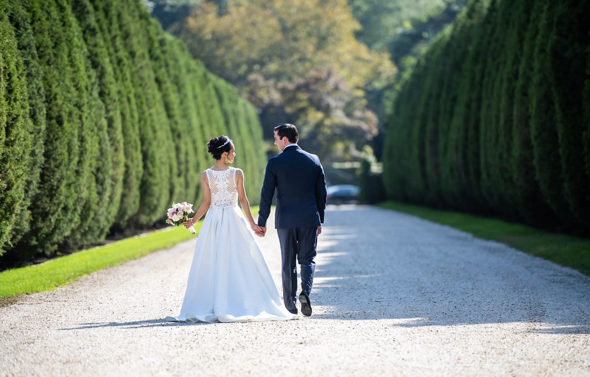 A bride and groom walking hand in hand on a gravel path surrounded by tall green hedges.