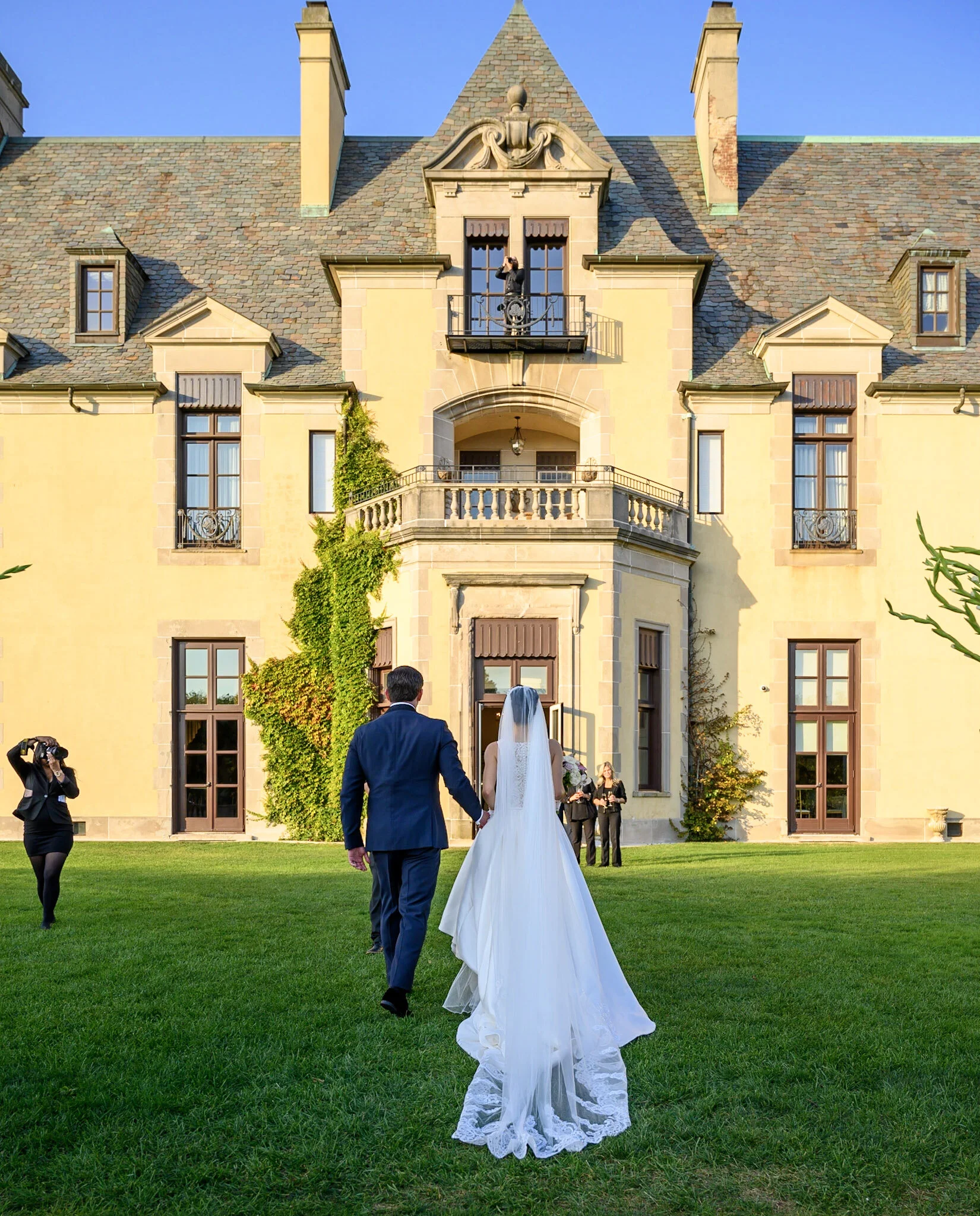 A bride and groom walking on a green lawn toward a large, elegant mansion with multiple windows, chimneys, and decorative architectural details, during daytime. The bride wears a white wedding gown with a veil, and the groom wears a dark suit. There 