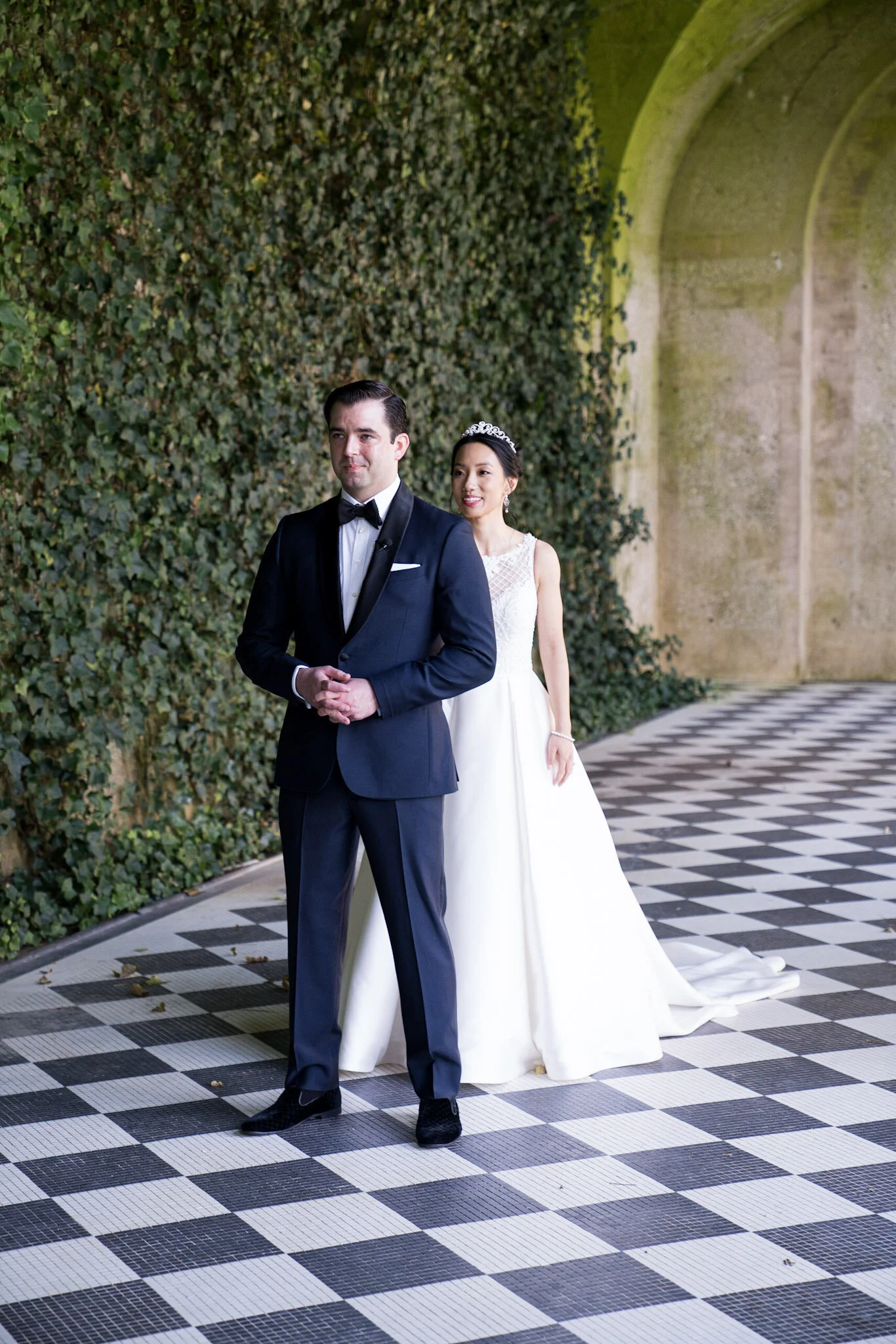 A groom in a tuxedo and a bride in a white wedding dress and tiara standing in front of a green ivy-covered wall with archways and a black-and-white checkered floor.