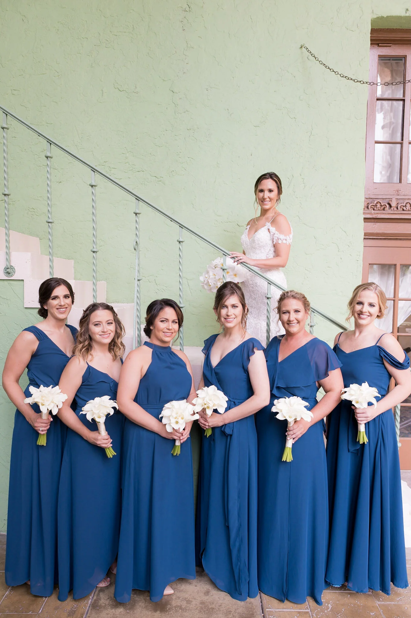 Bridal party of nine women, with the bride standing on a staircase holding a bouquet, and eight women in matching blue bridesmaids dresses holding white flowers, posed in front of a green textured wall with a window and a chain hanging from the ceili