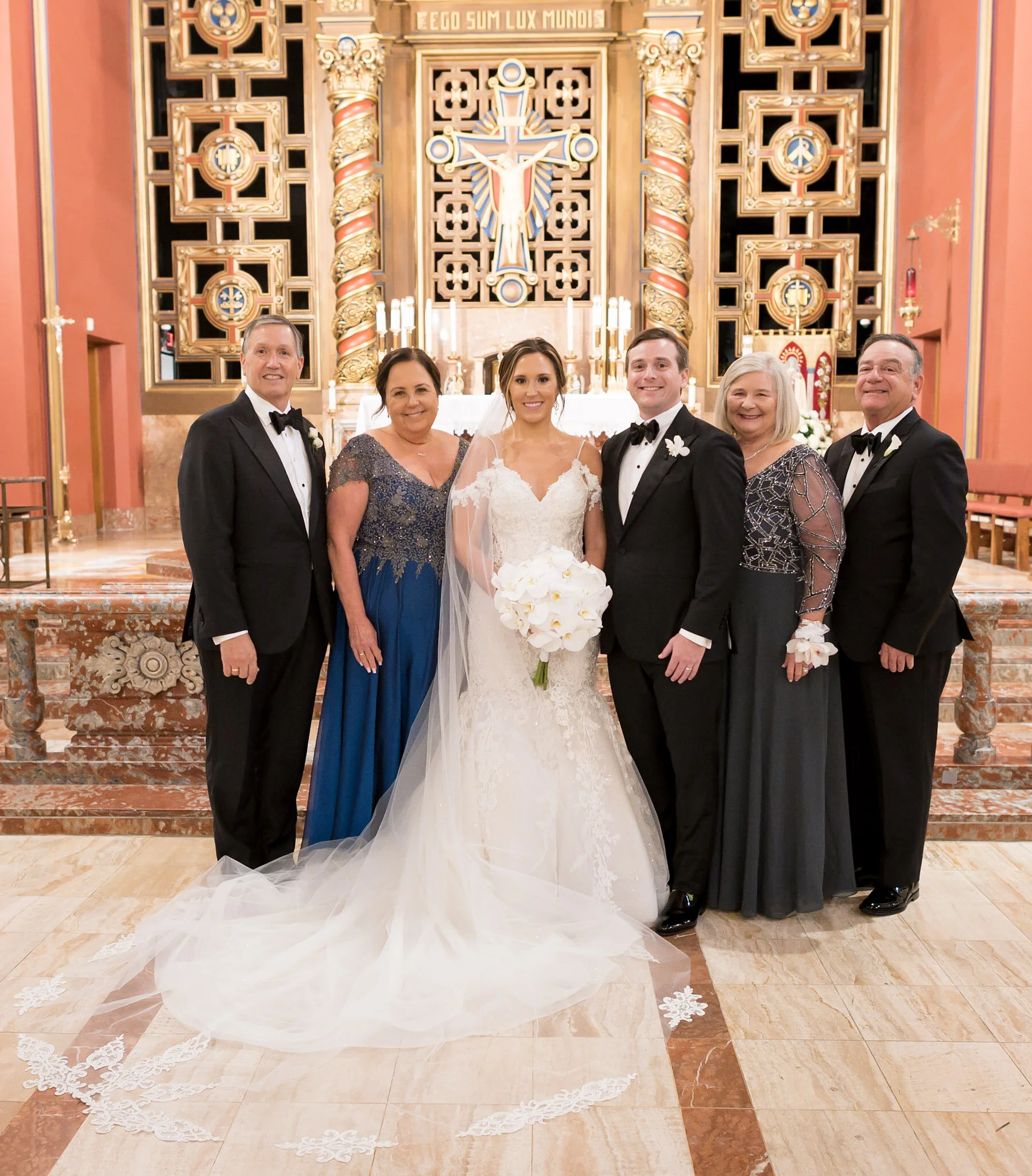 A wedding group photo inside a church with ornate decorations. The bride is in the center wearing a white wedding gown with a veil, surrounded by four men in tuxedos and two women in formal dresses. The backdrop features a large crucifix and religiou