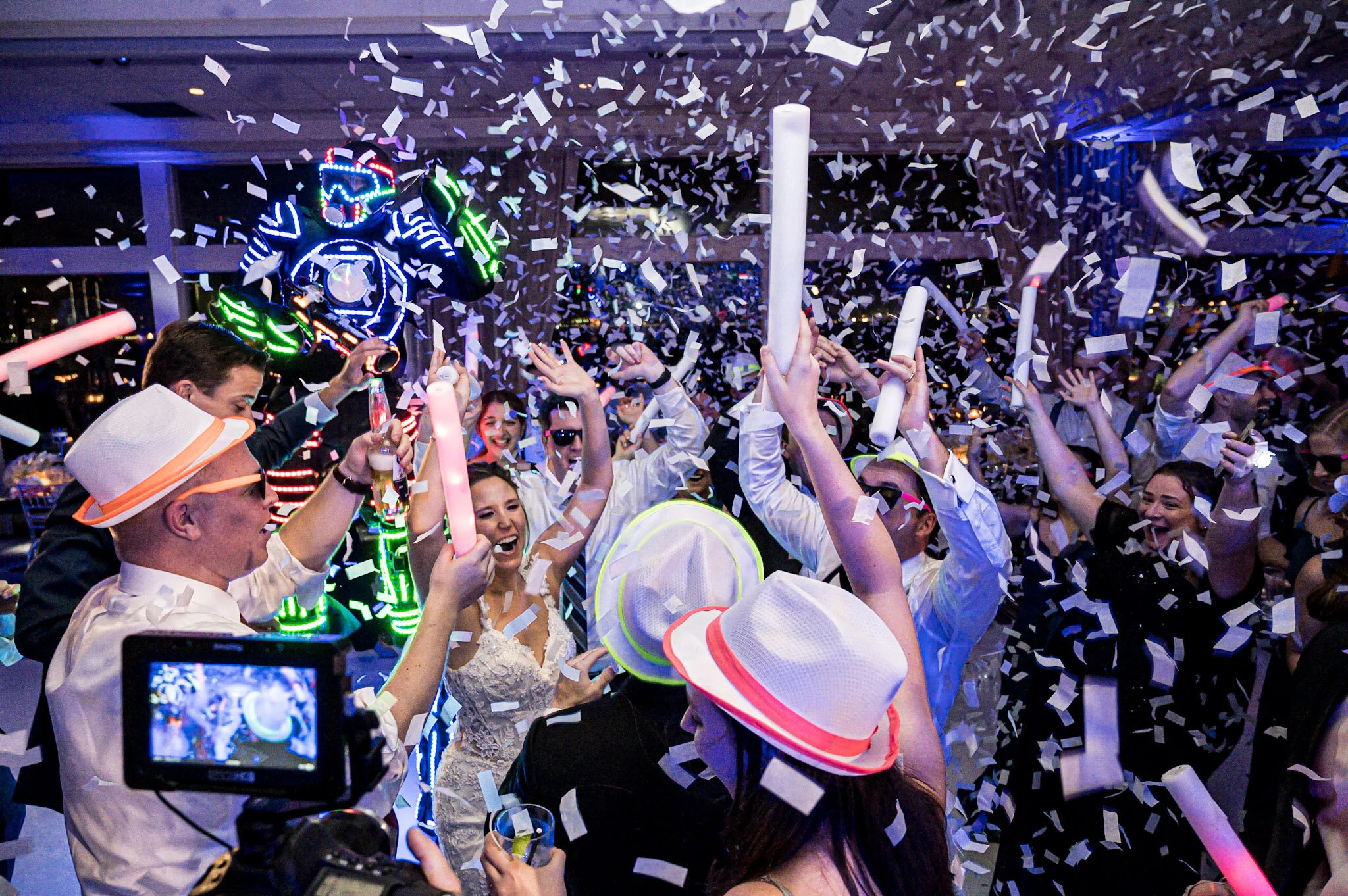 People celebrating at a party, some wearing white hats, surrounded by falling white confetti, with a person in a robot costume lit with colorful LED lights.
