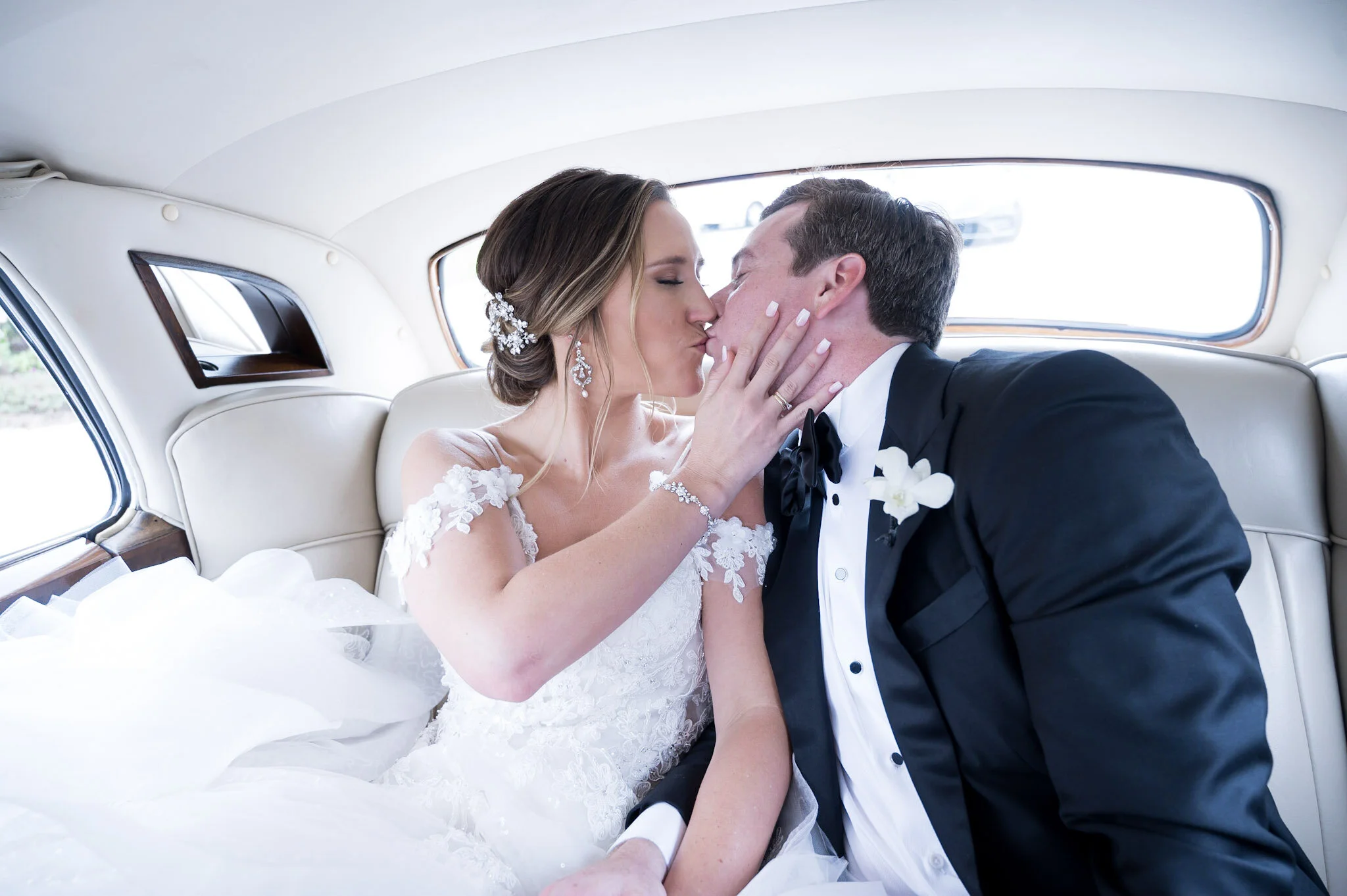 A bride and groom sharing a kiss inside a vintage car, with the bride wearing a white wedding dress and the groom in a black tuxedo.