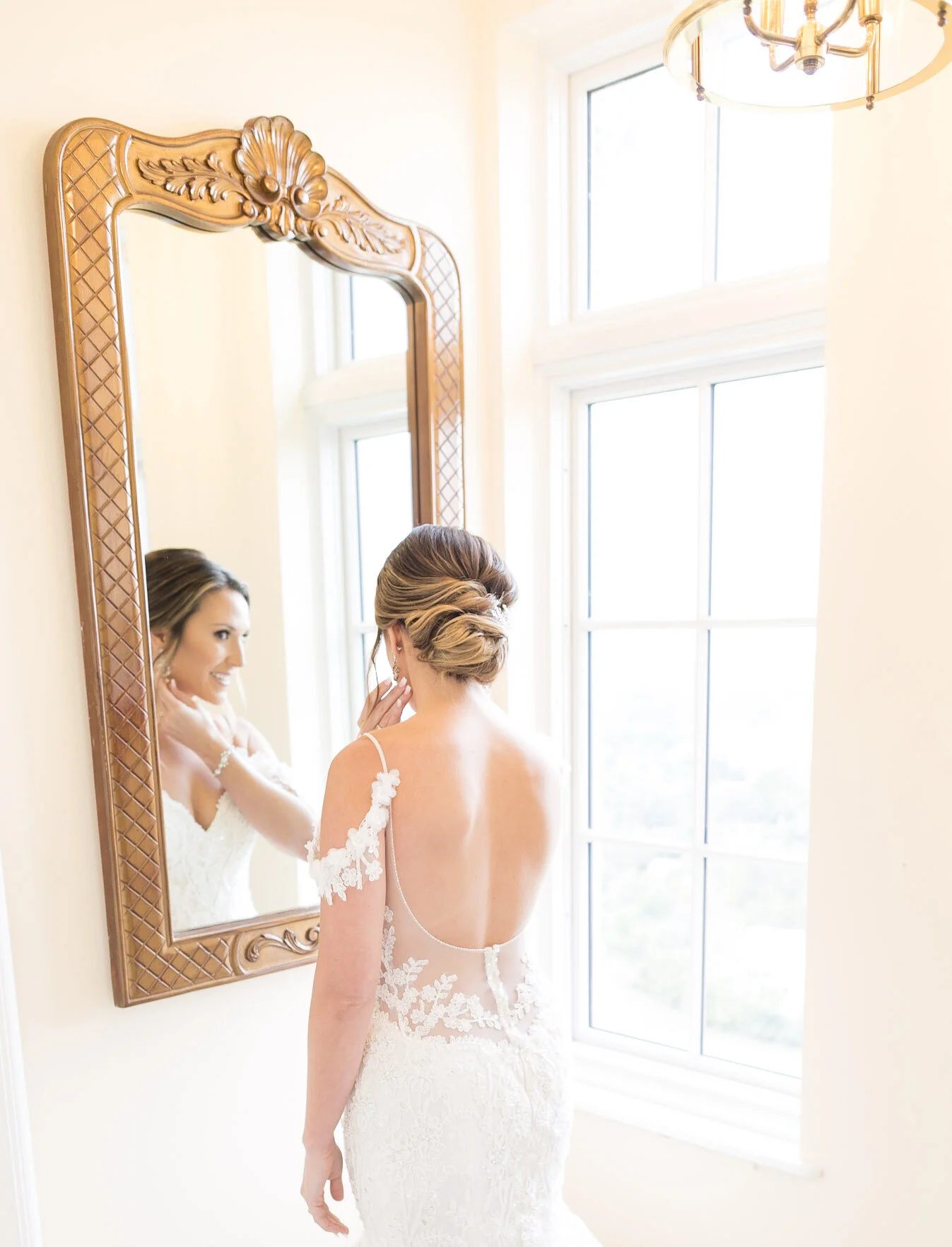 A bride looking at herself in a rectangular mirror with an ornate wooden frame as she gets ready, standing near a large window with natural light streaming in.