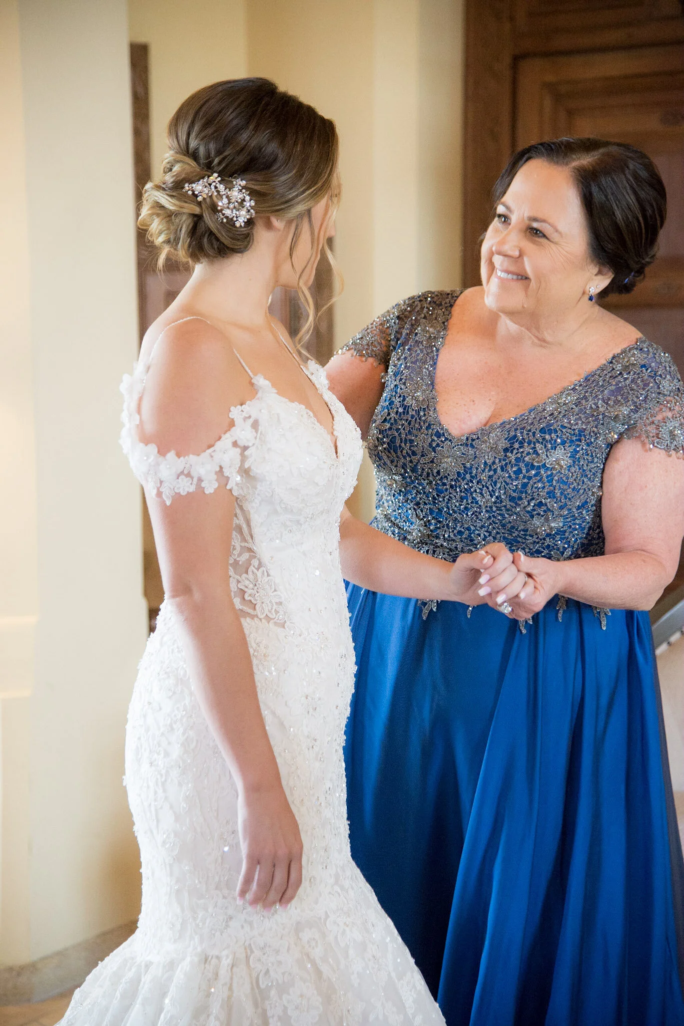 A bride in a white wedding dress shaking hands with a woman in a blue and silver dress, smiling at each other.