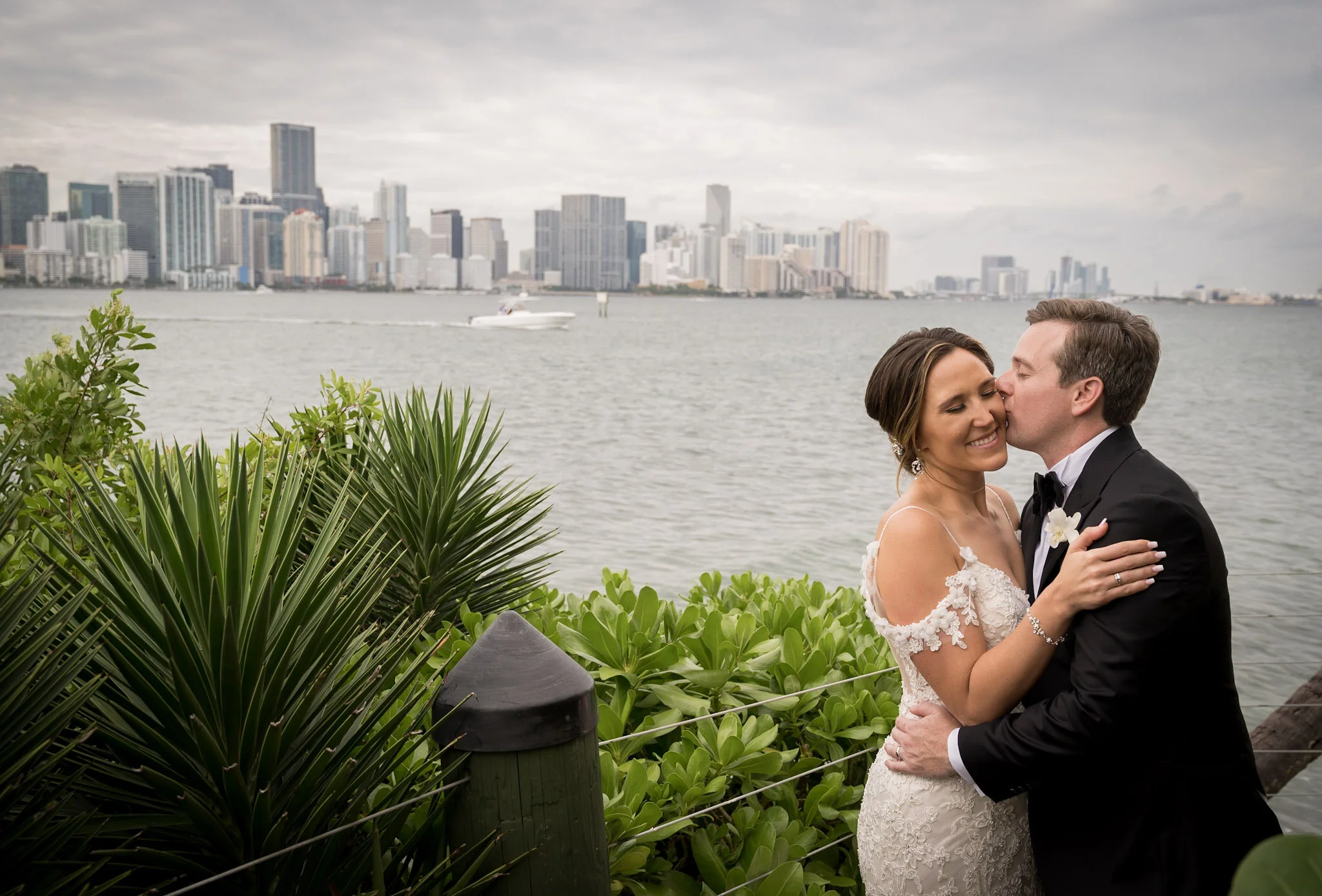 A newlywed couple in wedding attire sharing a romantic moment by a waterfront with city skyline in the background.