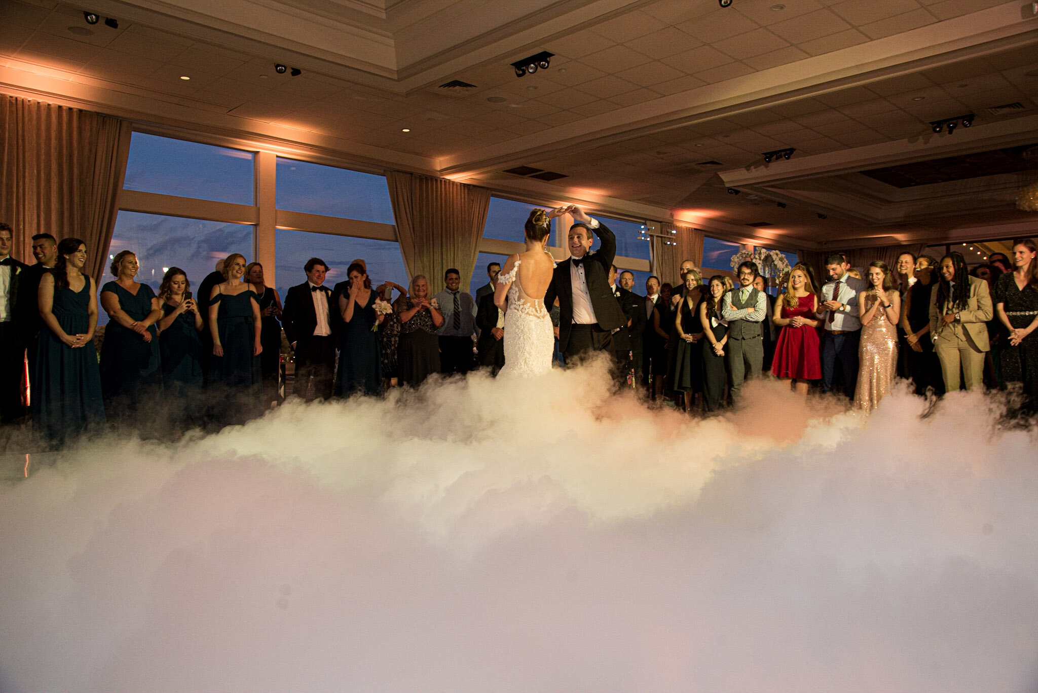 A bride and groom sharing their first dance at a wedding reception, surrounded by guests, with a fog machine creating a dreamy atmosphere, in a room with large windows showing the night sky.