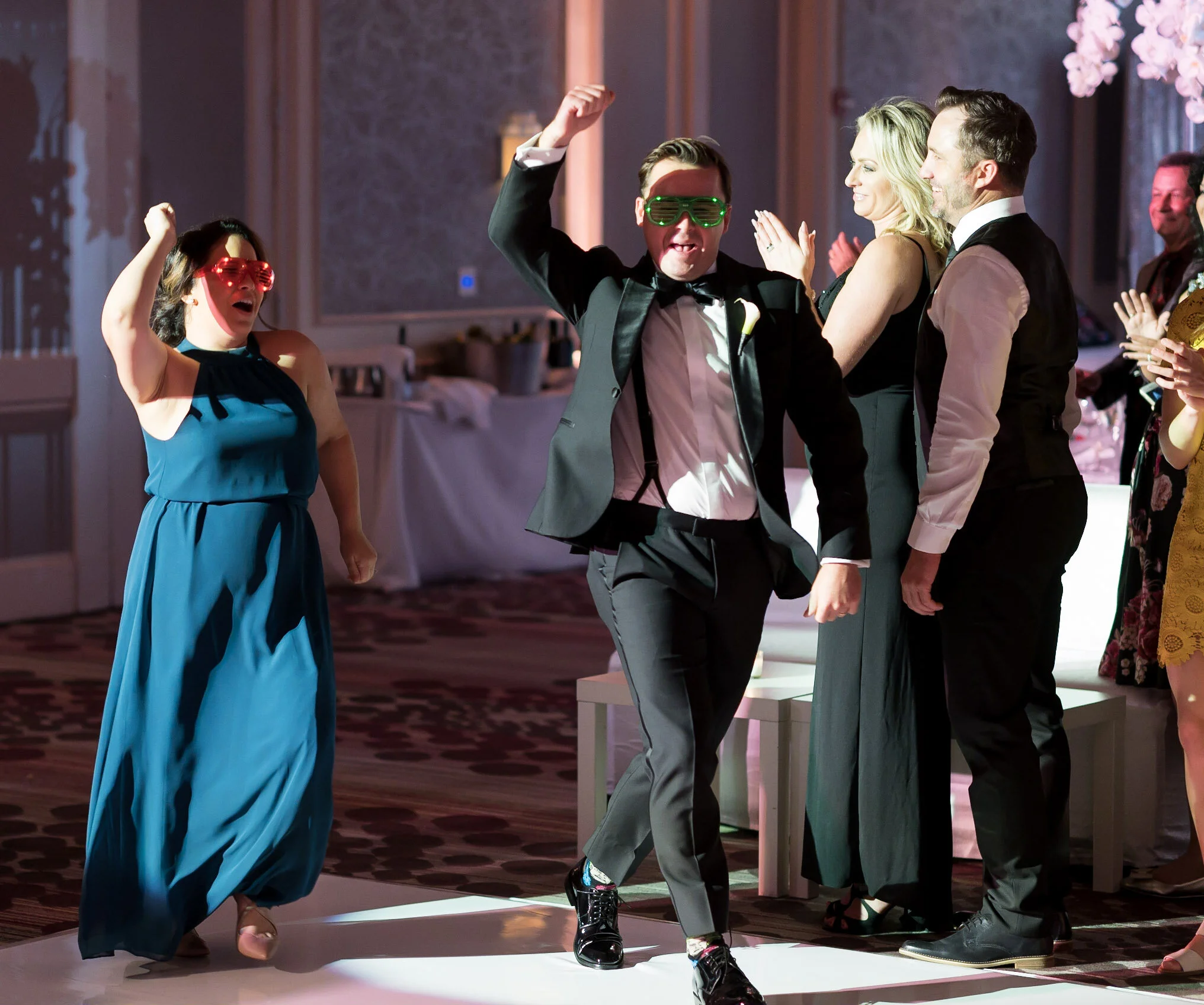 People dancing at a celebration, some wearing colorful glasses, in a room with ornate decor.
