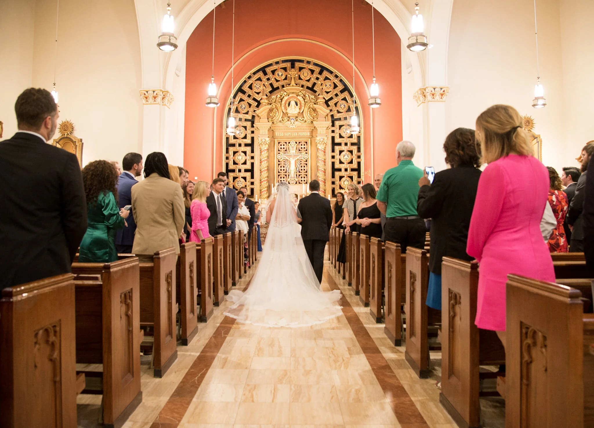 A wedding ceremony inside a church with guests standing along the aisle. The bride and groom are at the altar in front of a decorative gold and black religious altar, with a veil trailing down the aisle behind them. The church has high arched ceiling