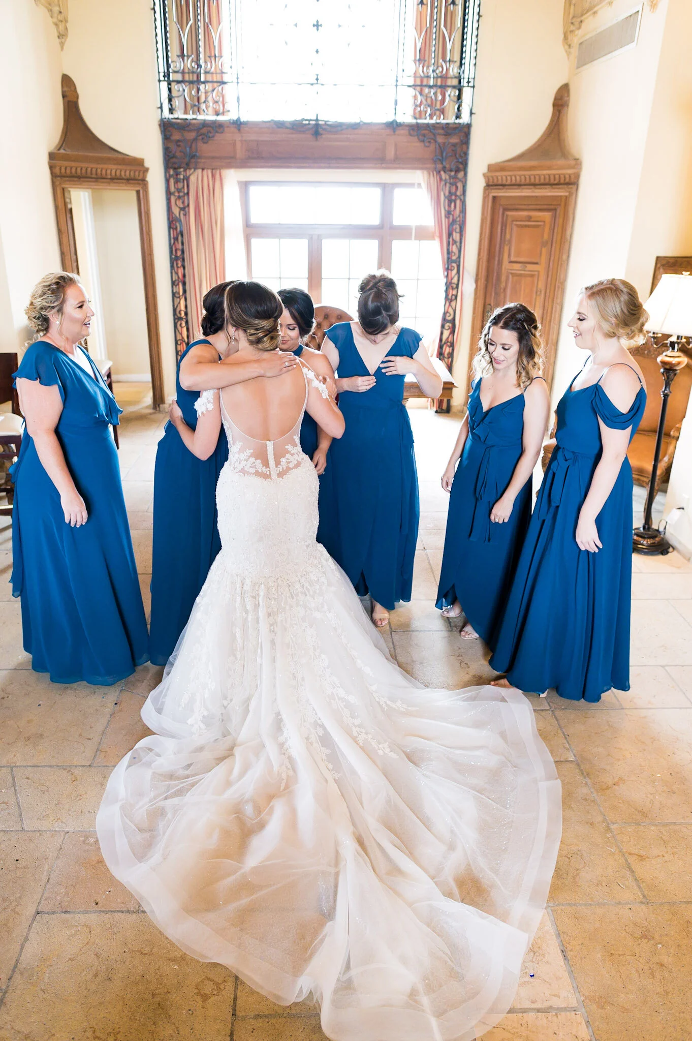A bride in a white lace wedding gown with a long train receiving help from her bridesmaids, dressed in blue dresses, in a well-lit room with large windows and ornate woodwork.
