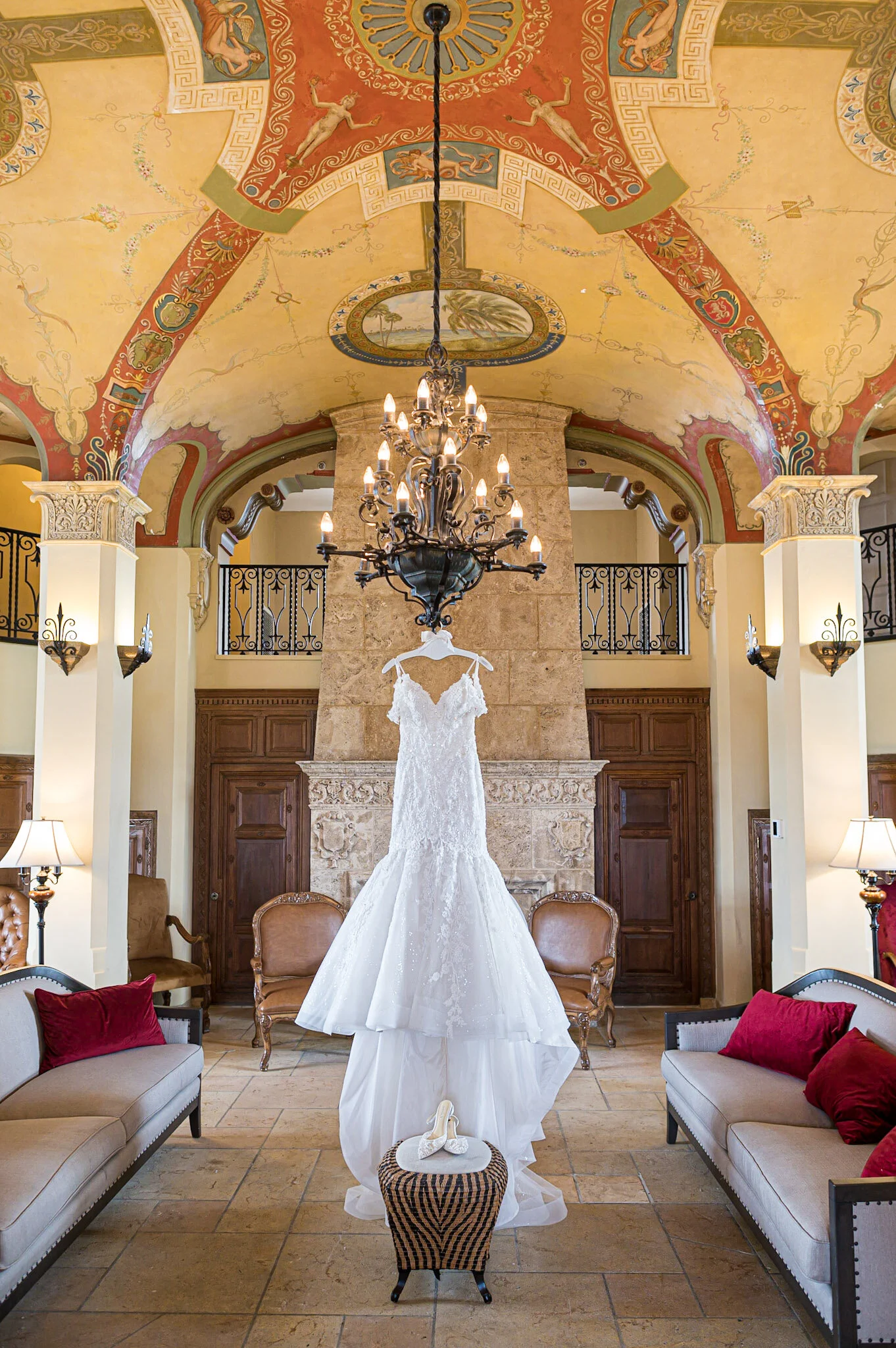 A wedding dress hanging in a luxurious room with a vibrant, ornate ceiling. The room features a chandelier, armchairs with red pillows, and decorative lighting fixtures.