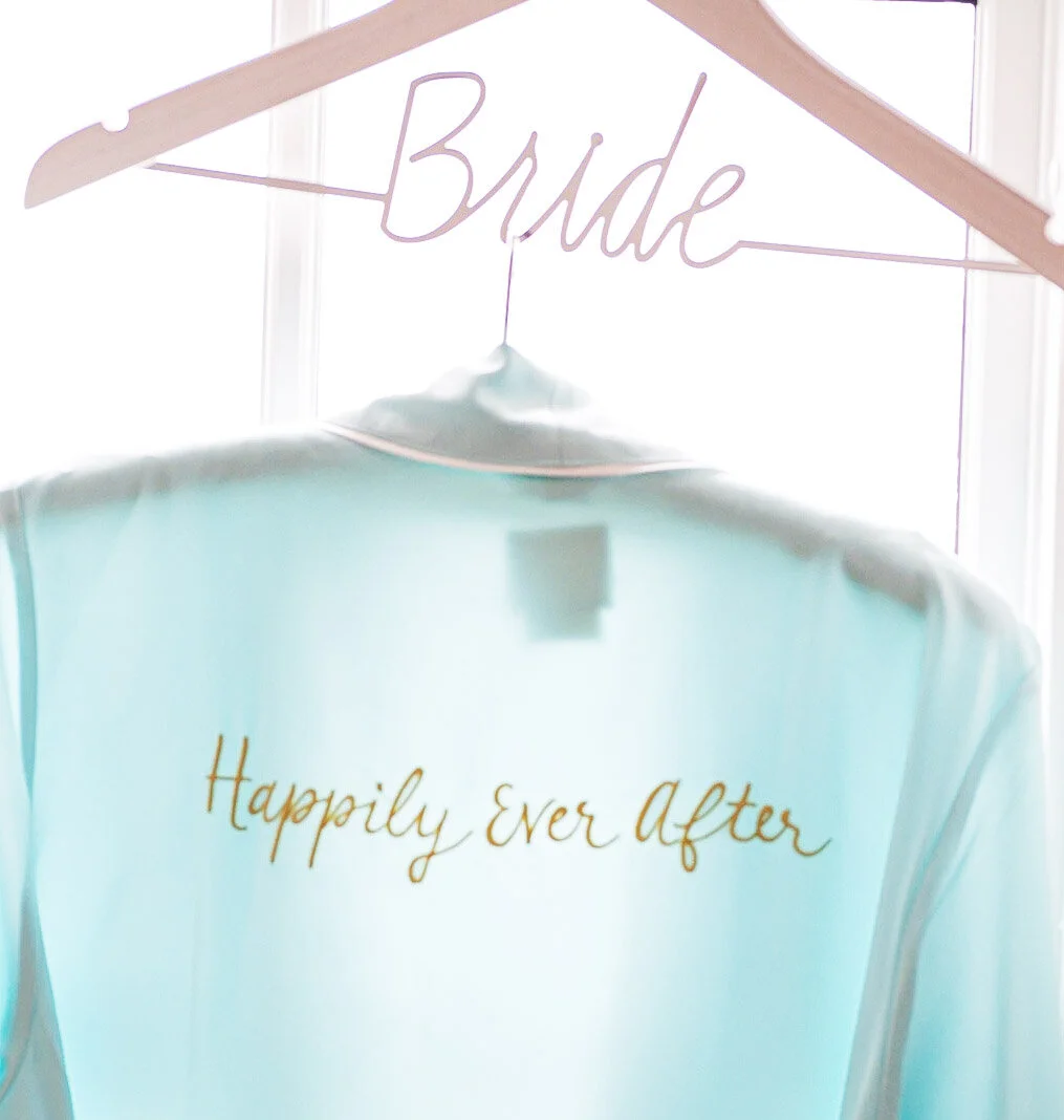 A light-colored wedding dress with the words "Happily Ever After" embroidered on the back. The dress is hanging on a pink hanger with a sign that says "Bride."