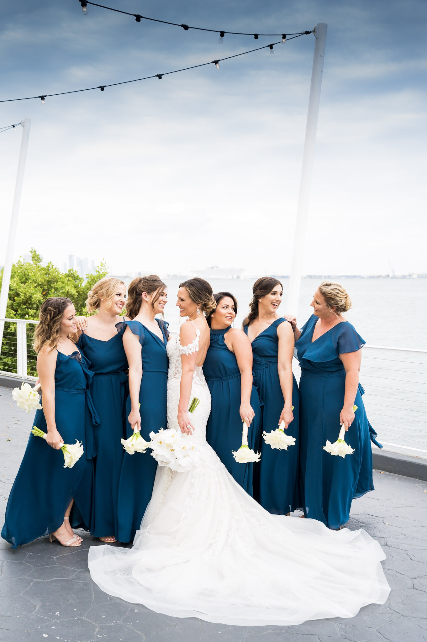 A group of eight women in matching blue bridesmaid dresses and a bride in a white wedding gown with a train, holding white flower bouquets, gathered outdoors near water with cityscape in the background, during daytime.
