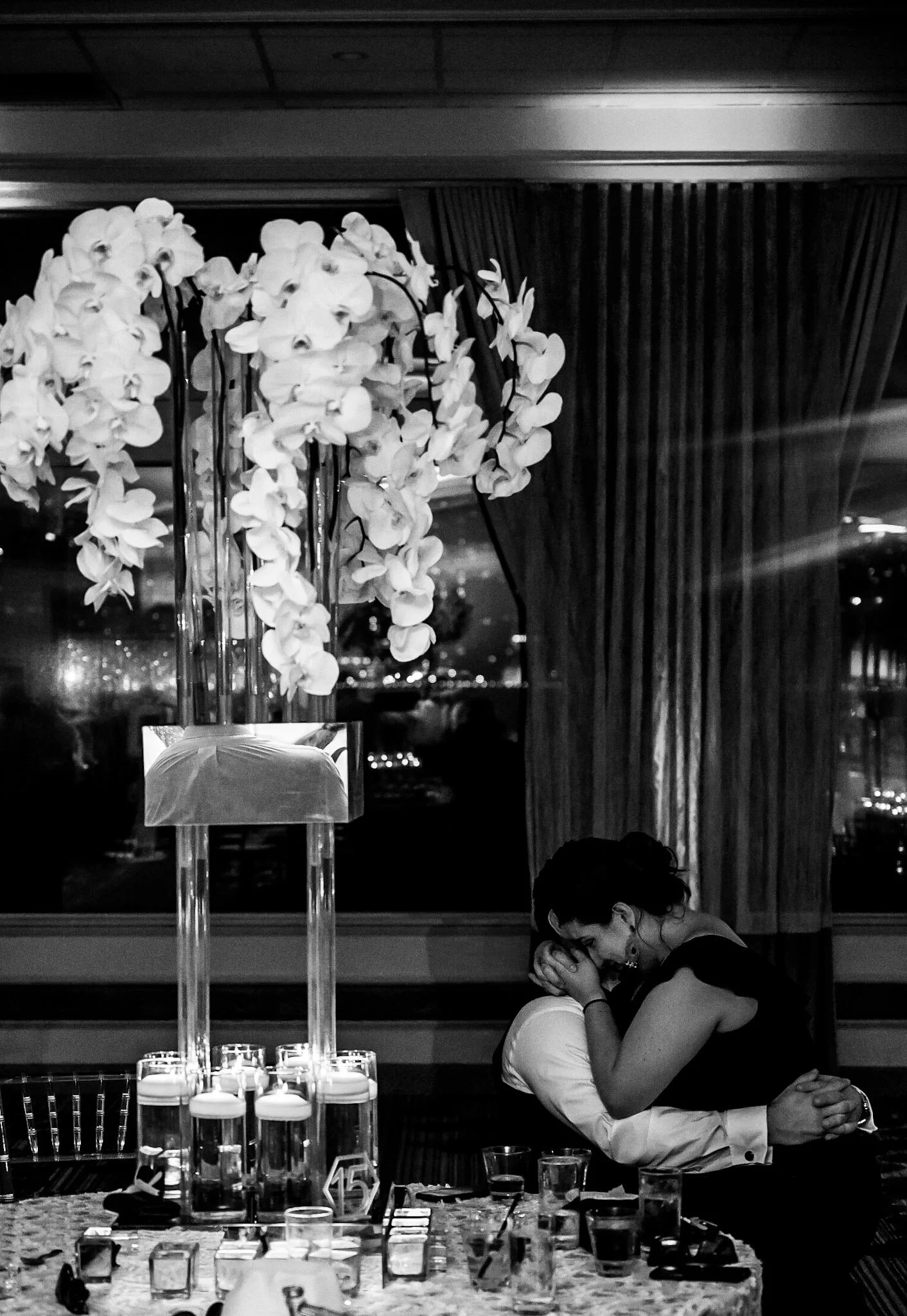 A woman in formal attire with clasped hands leaning on a table at a formal event. In the background, there is a tall floral arrangement with white orchids, and city lights are visible through window curtains.
