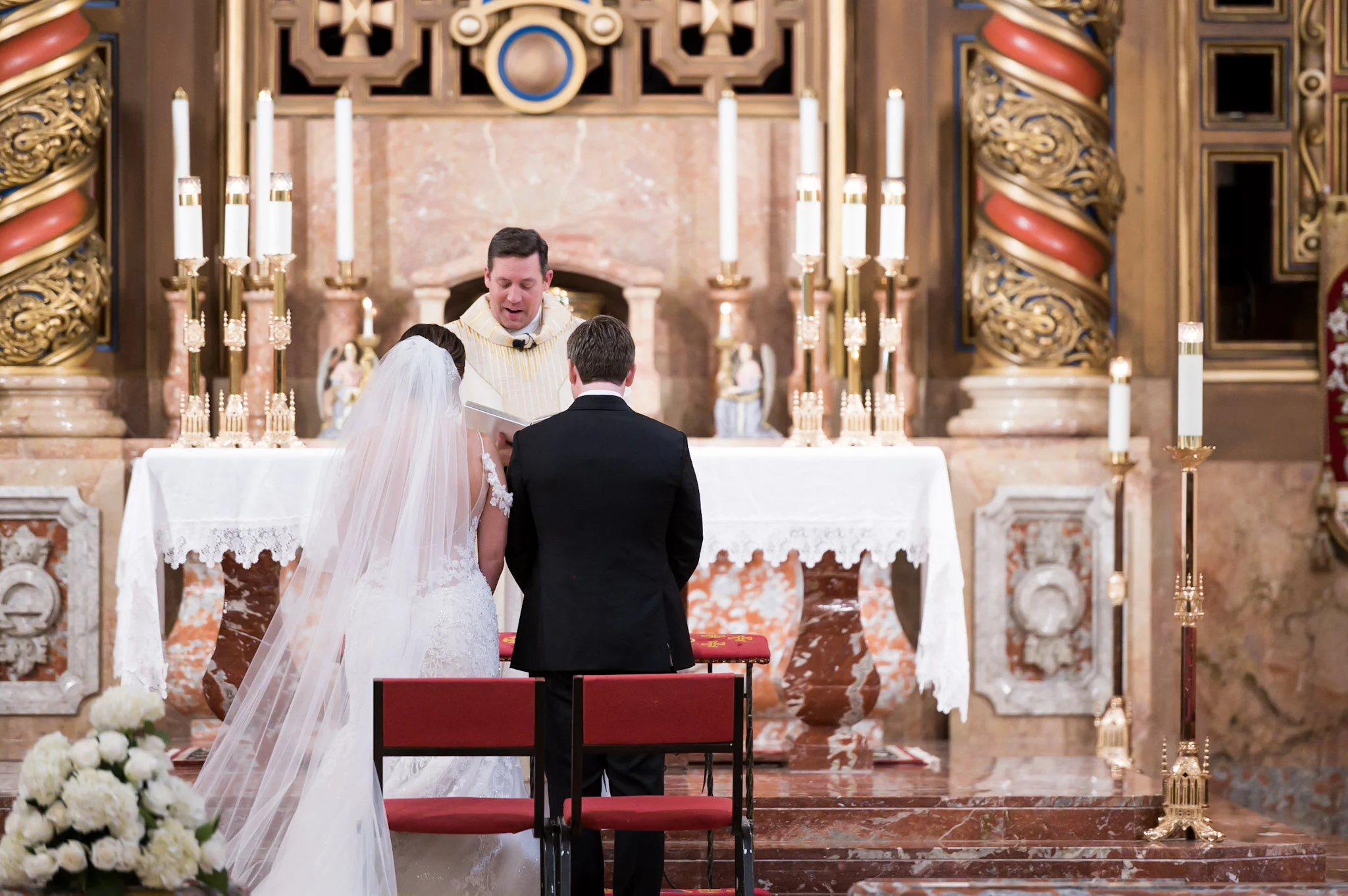 A bride and groom standing before a priest in a church during their wedding ceremony, with ornate decorations and candles in the background.