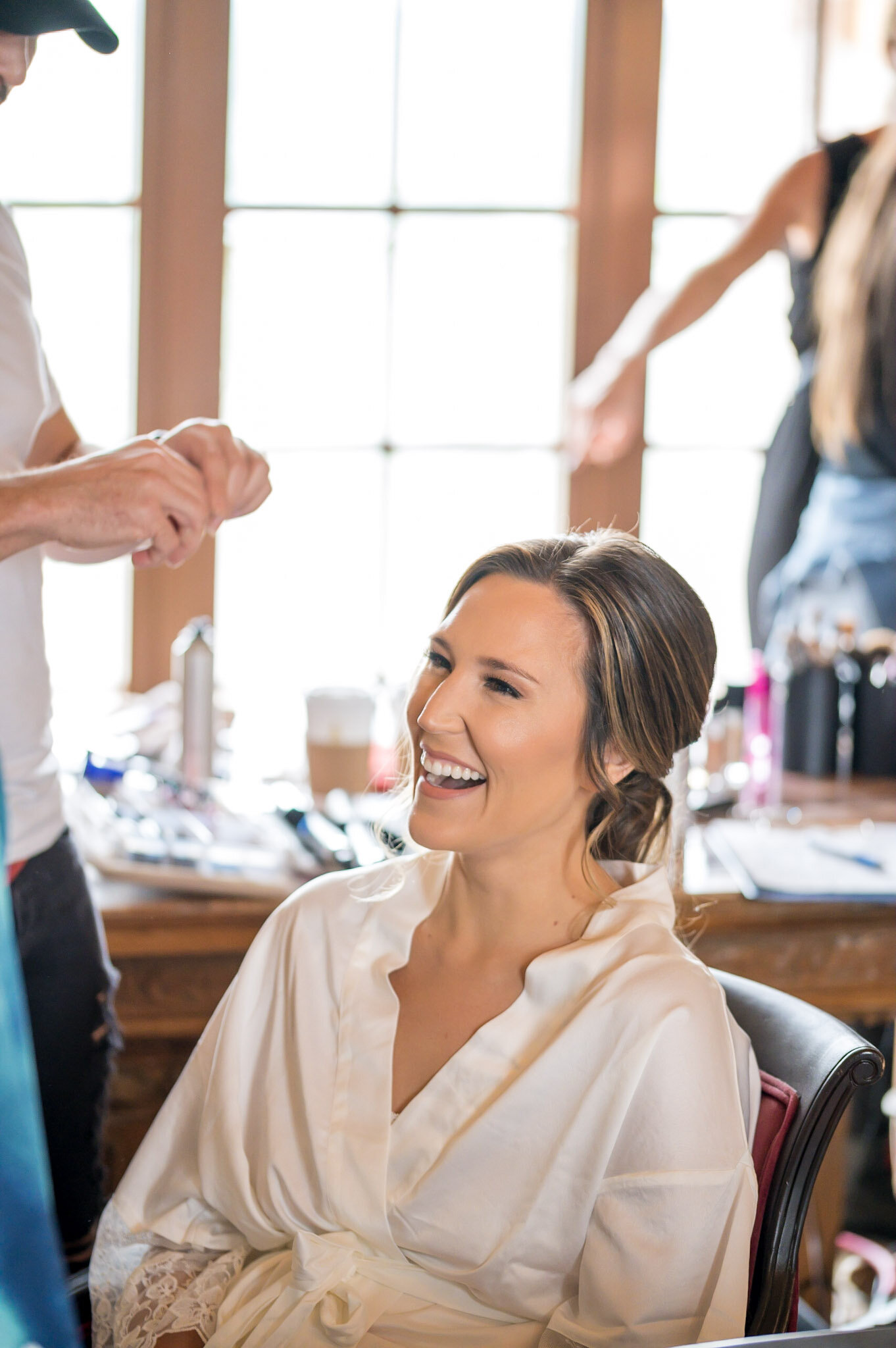 A woman sitting in a salon chair with a cheerful expression as a hair stylist works on her hair, with salon tools and large windows in the background.