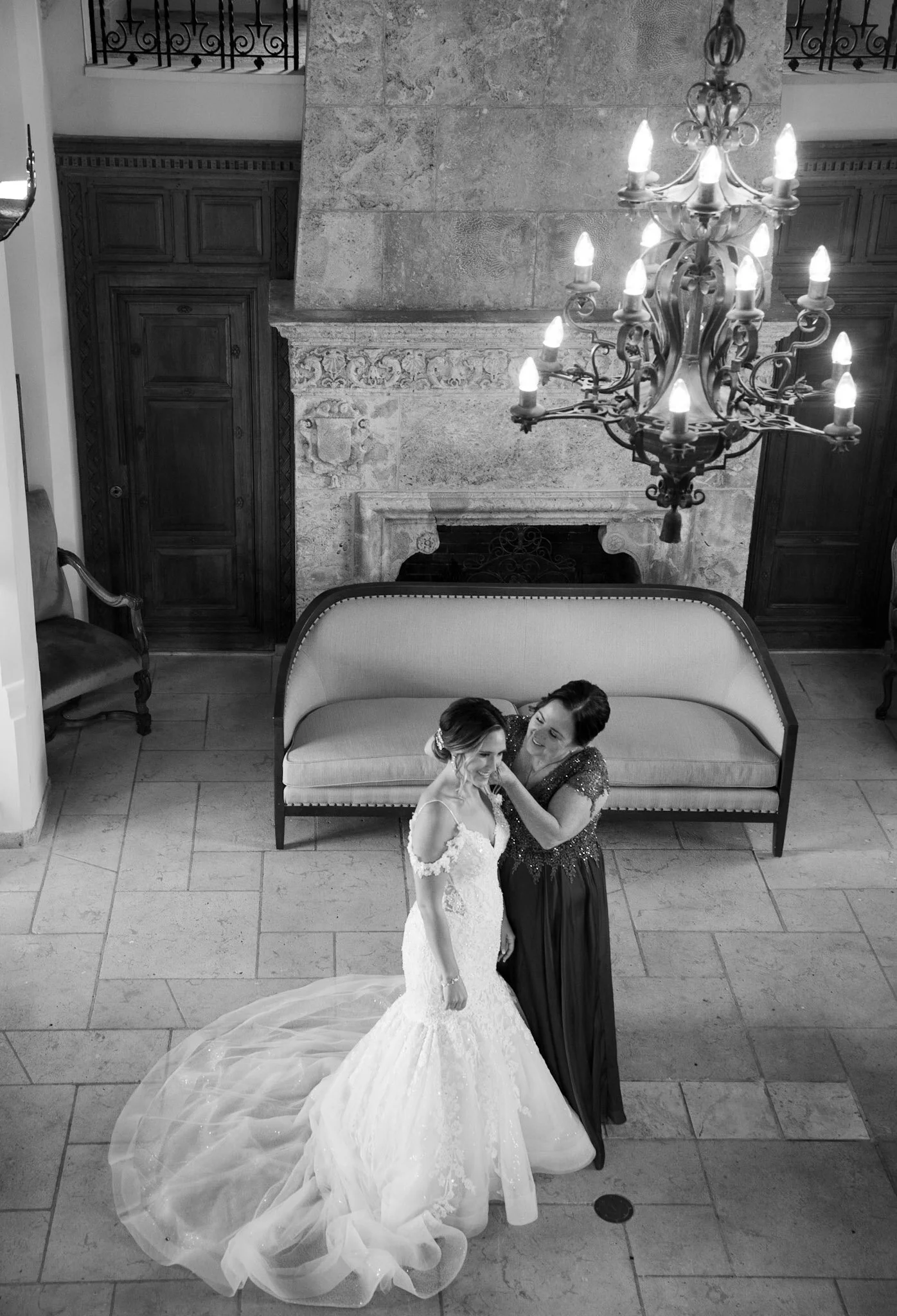 Two women at a wedding reception, one in a wedding dress and the other in a formal dress, smiling and embracing under a chandelier in a room with stone fireplace and wooden cabinets.