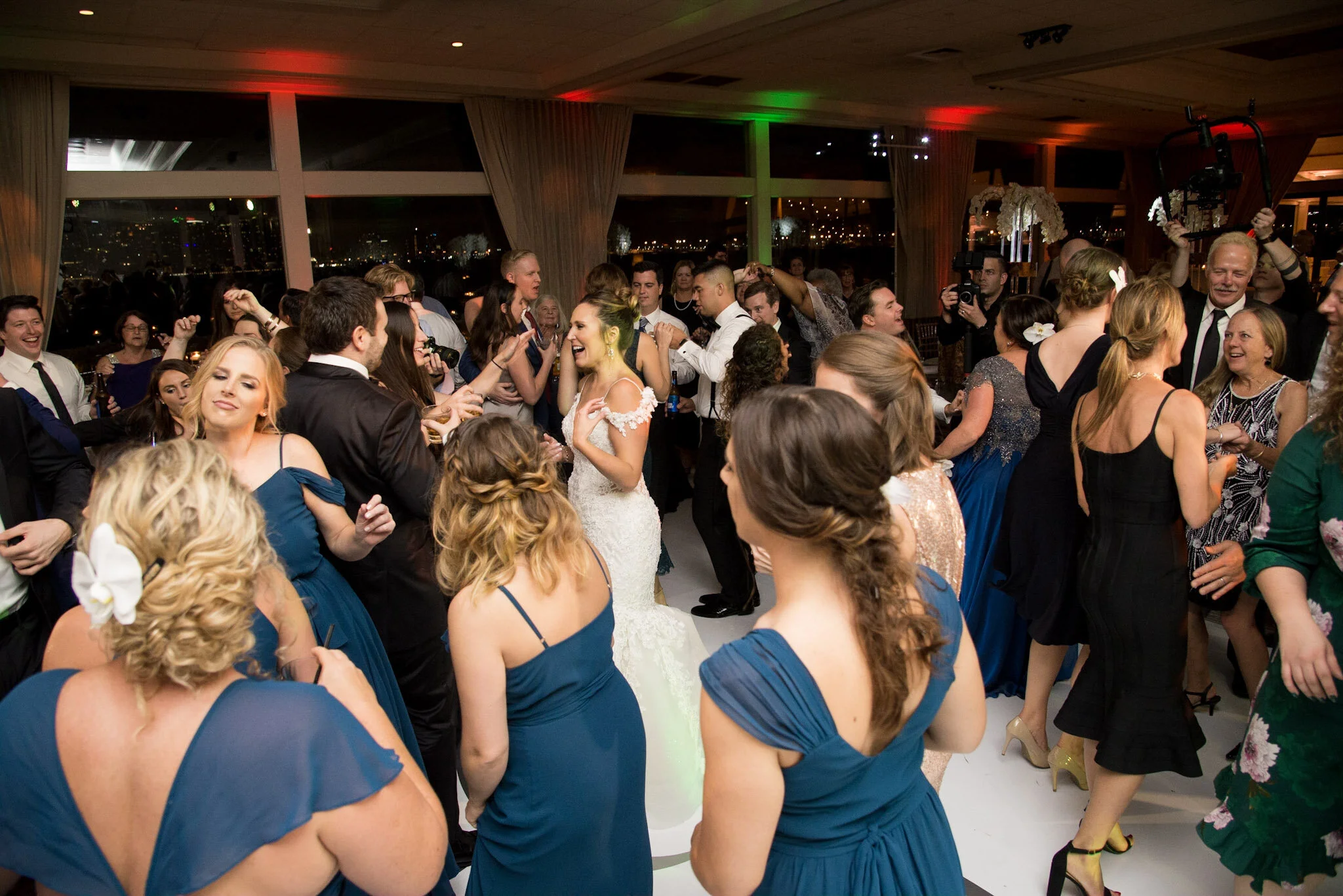 Celebration party with people dancing and socializing indoors, with city lights visible through large windows at night.