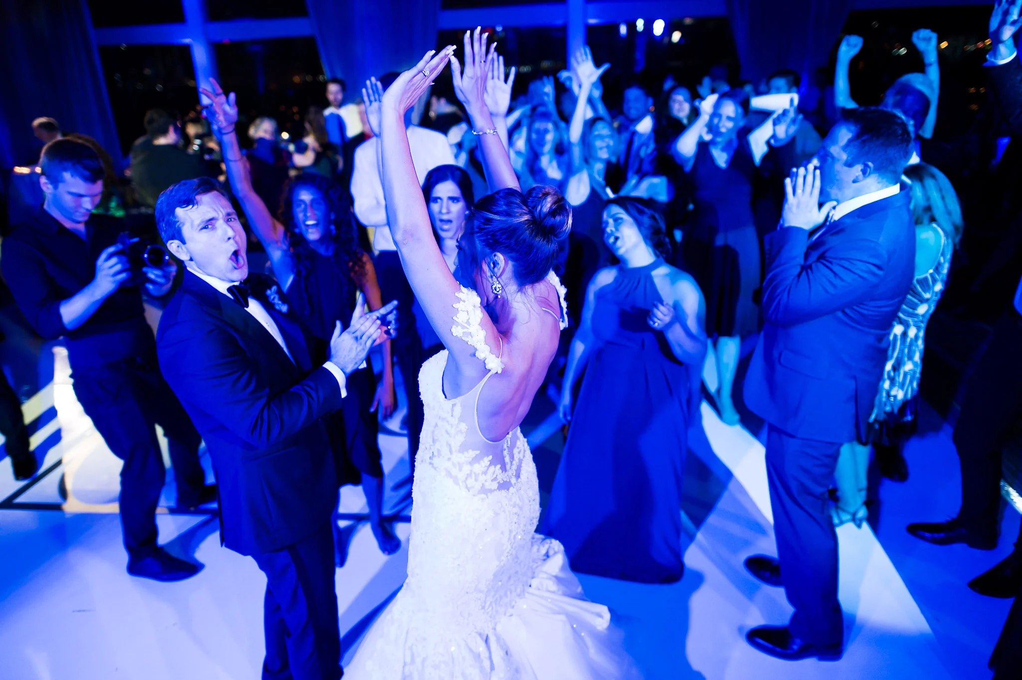 People dancing at a wedding reception with blue lighting, with a bride wearing a white lace gown in the center raising her hands.