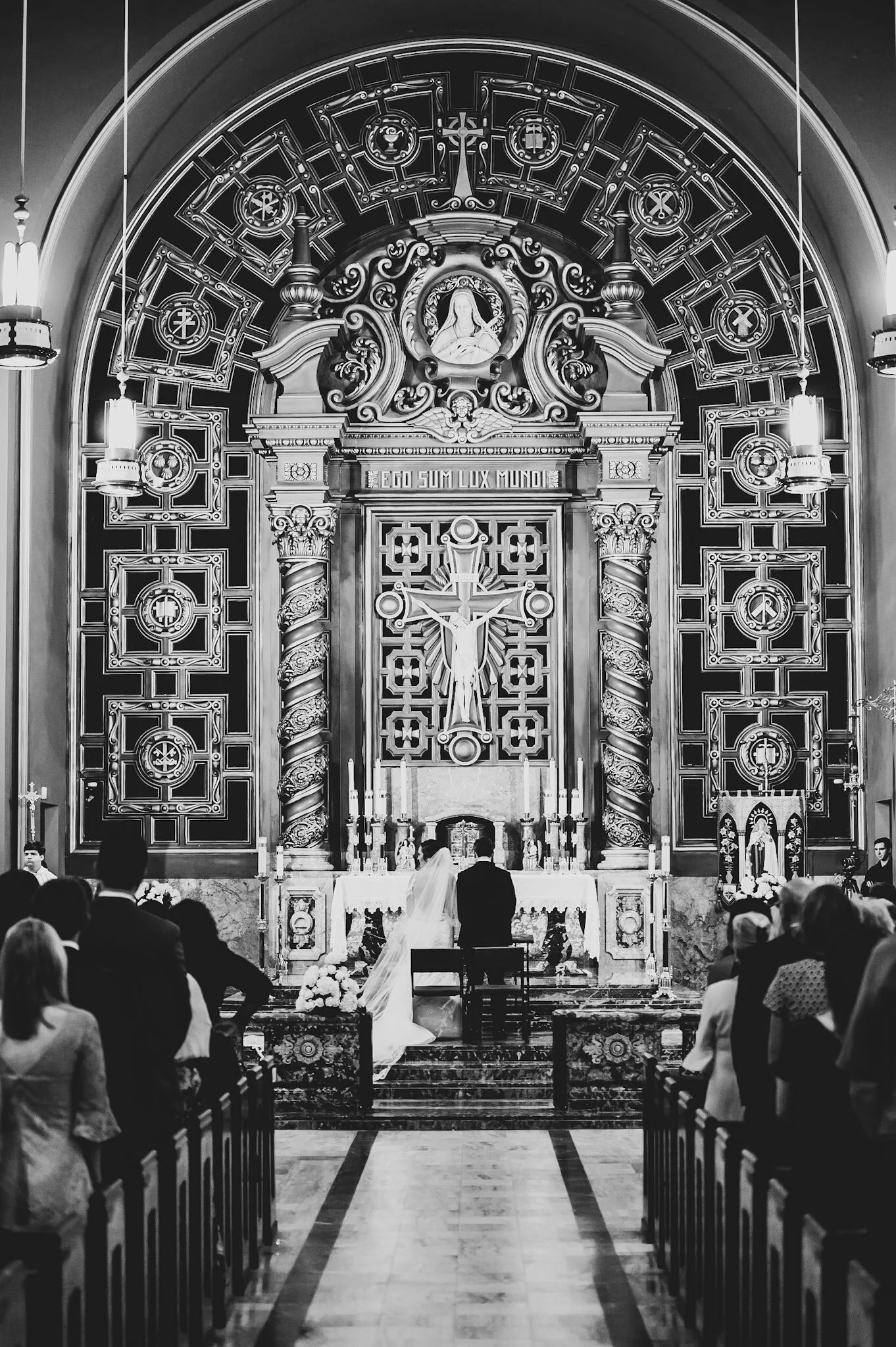A black and white photo of a church altar during a wedding ceremony, with a bride and groom standing in front of the priest. The altar is ornate with religious symbols, candles, and a crucifix.