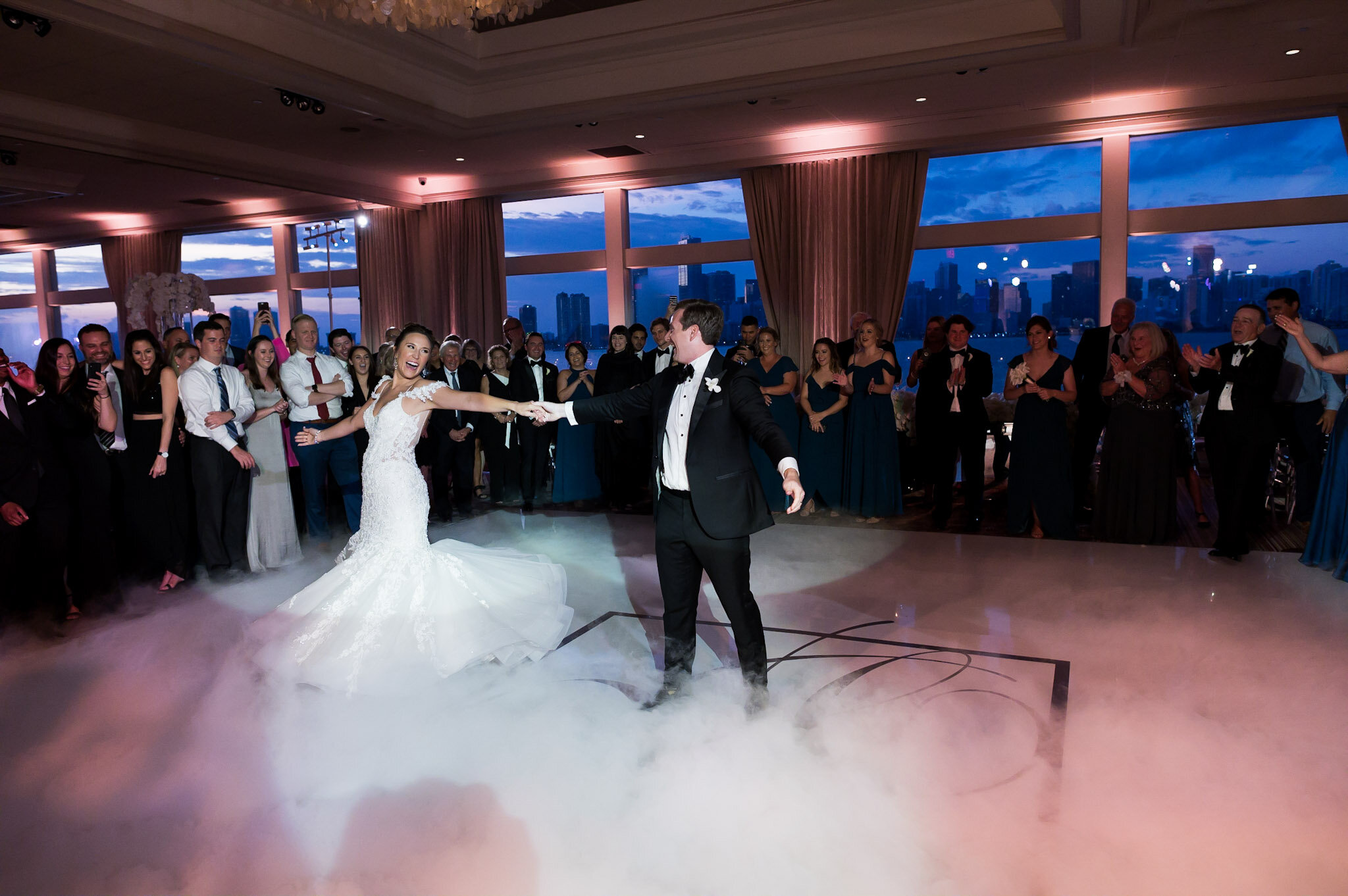 A wedding reception with a bride and groom dancing in front of a crowd in a high-rise venue with city skyline views at dusk.