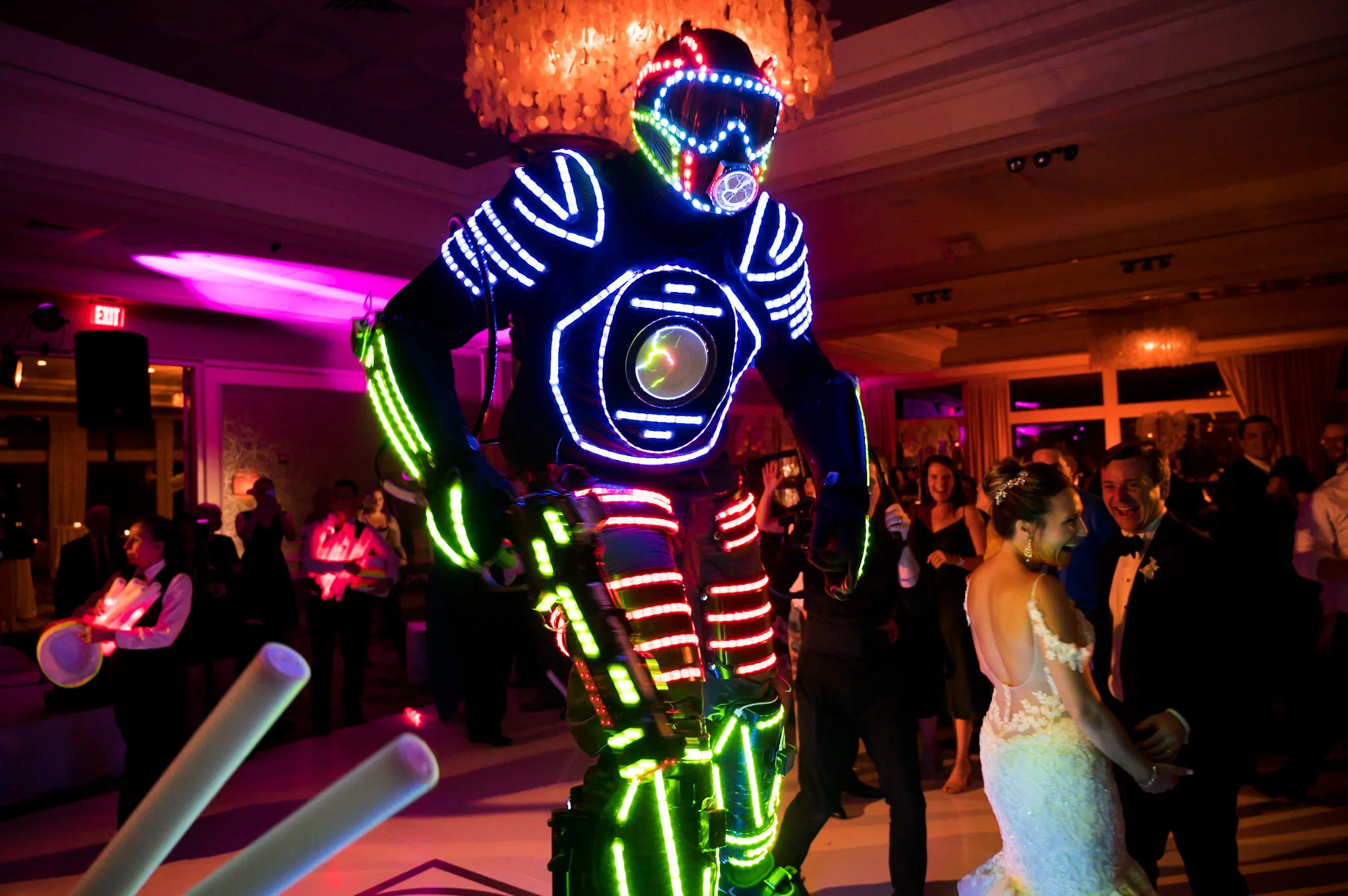 A performer dressed in a futuristic, LED-lit costume with a helmet and mask, entertaining guests at a celebration with colorful lights and a lively crowd dancing and smiling.