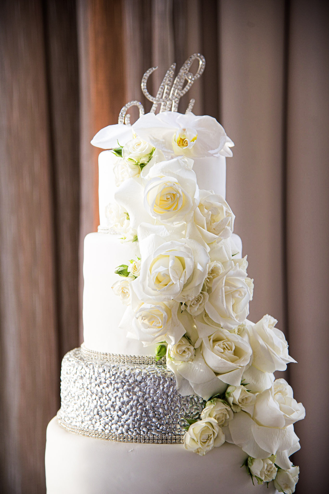 A tiered wedding cake decorated with white roses and orchids, with a shimmering silver band around the middle tier and a glittery cake topper that reads 'Love'.