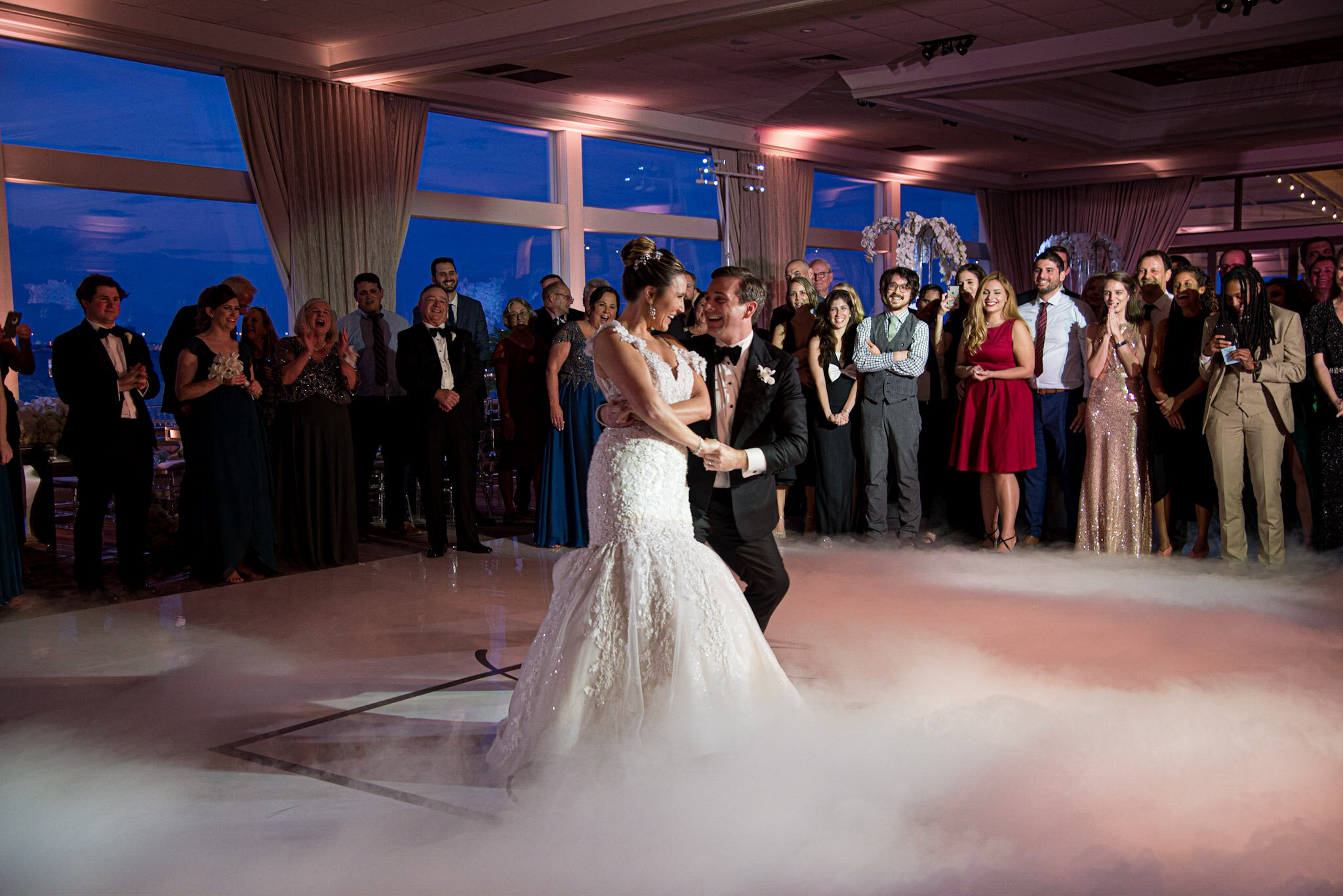A bride and groom dancing at their wedding reception surrounded by guests indoors with large windows showing a view of the city skyline at night.