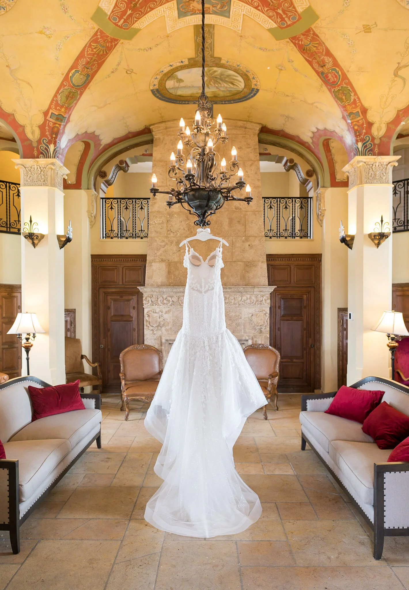 A wedding dress hangs from a chandelier in a decorated room with murals on the ceiling, stone fireplace, and sofas with red pillows.