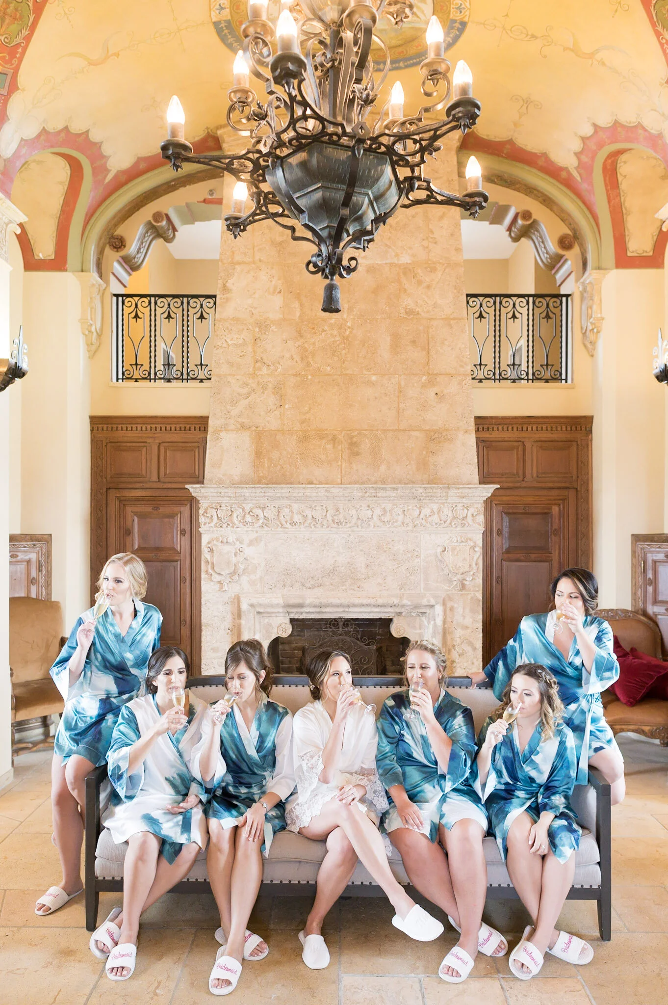 A group of women in matching robes sitting together in a living room, some drinking champagne, with two women in robes standing on each side.