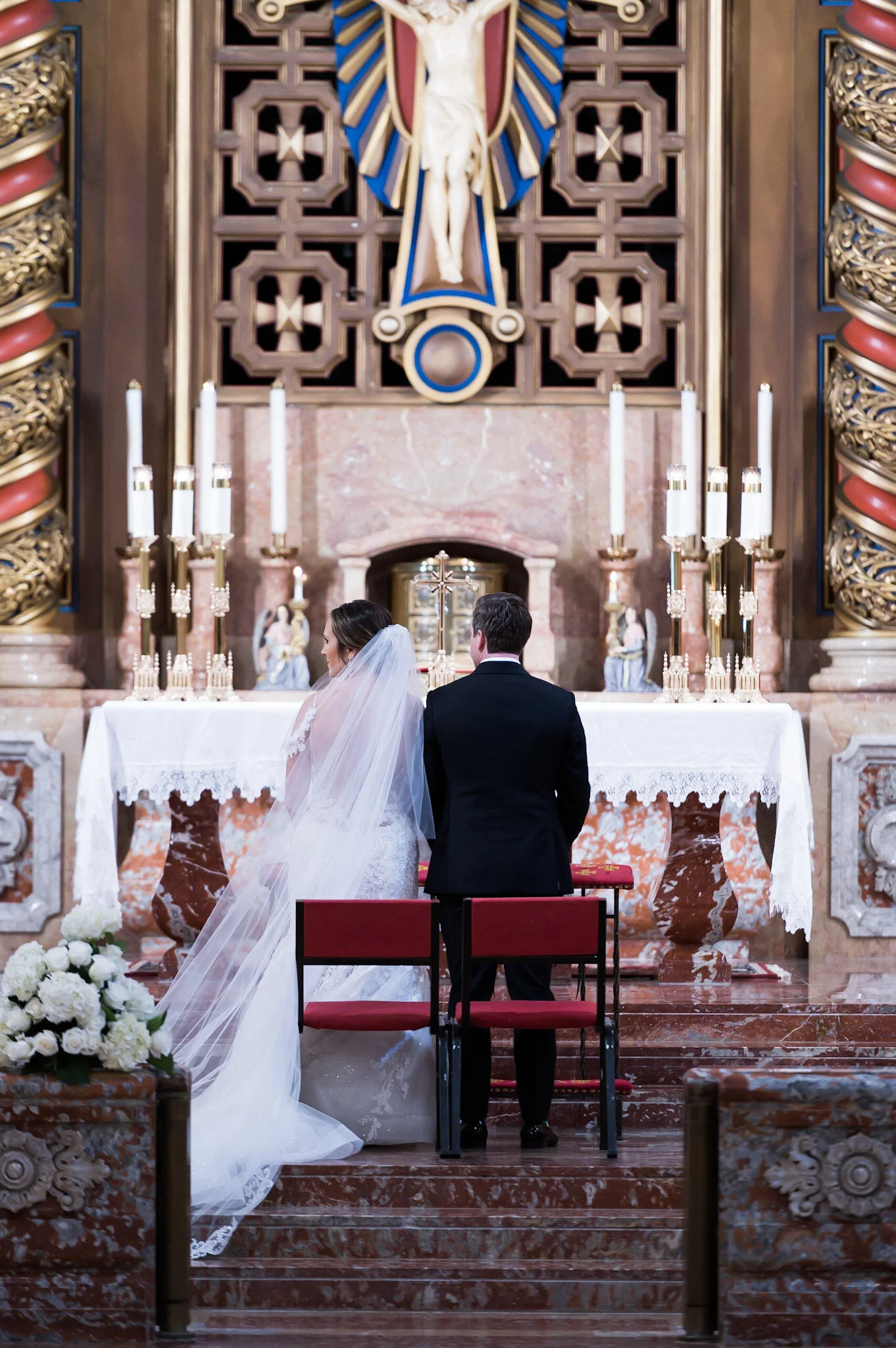 A bride and groom kneeling at an altar during a wedding ceremony in a church with ornate woodwork, gold accents, and religious symbols, including a crucifix and a large statue of Jesus Christ.