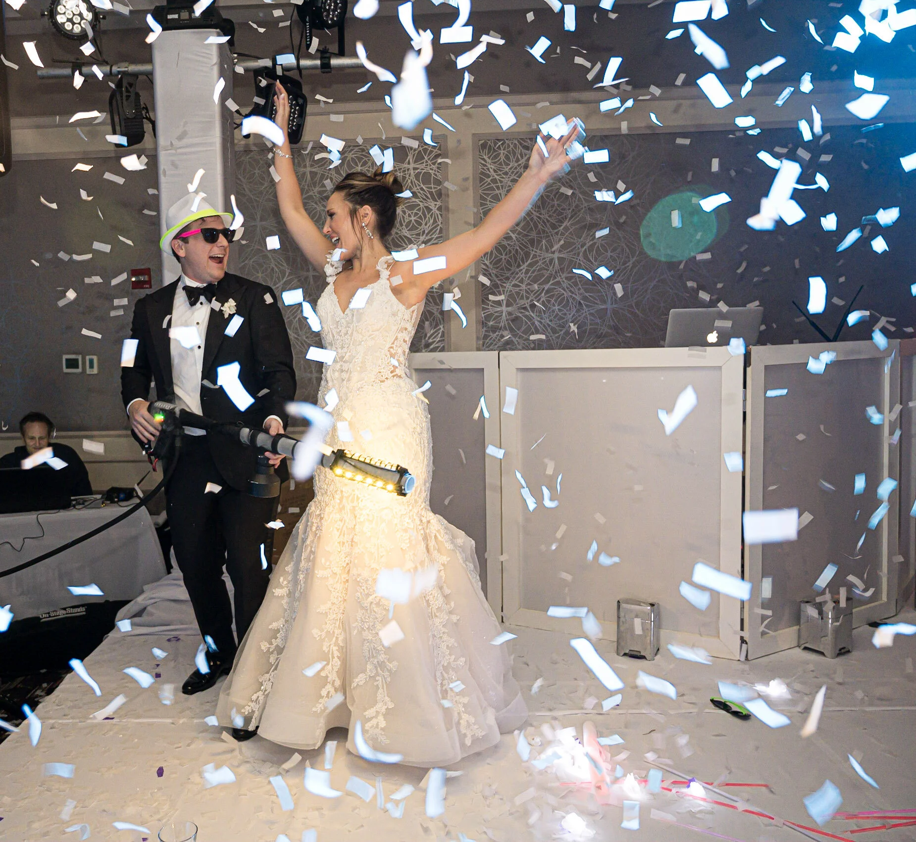 A bride and groom celebrating at their wedding reception, surrounded by falling confetti. The bride is wearing a white lace wedding dress with her arms raised, and the groom is dressed in a tuxedo with sunglasses and a hat, holding a microphone.
