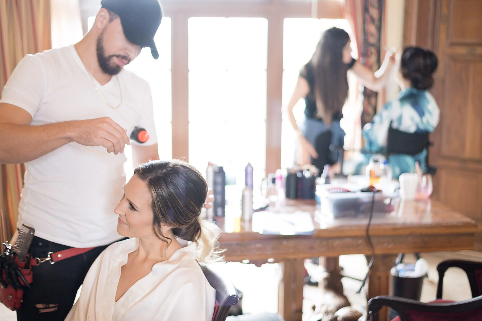 A hairstylist in a black cap and white shirt styles a woman with light brown hair in a salon. Other clients and stylist are visible in the background.