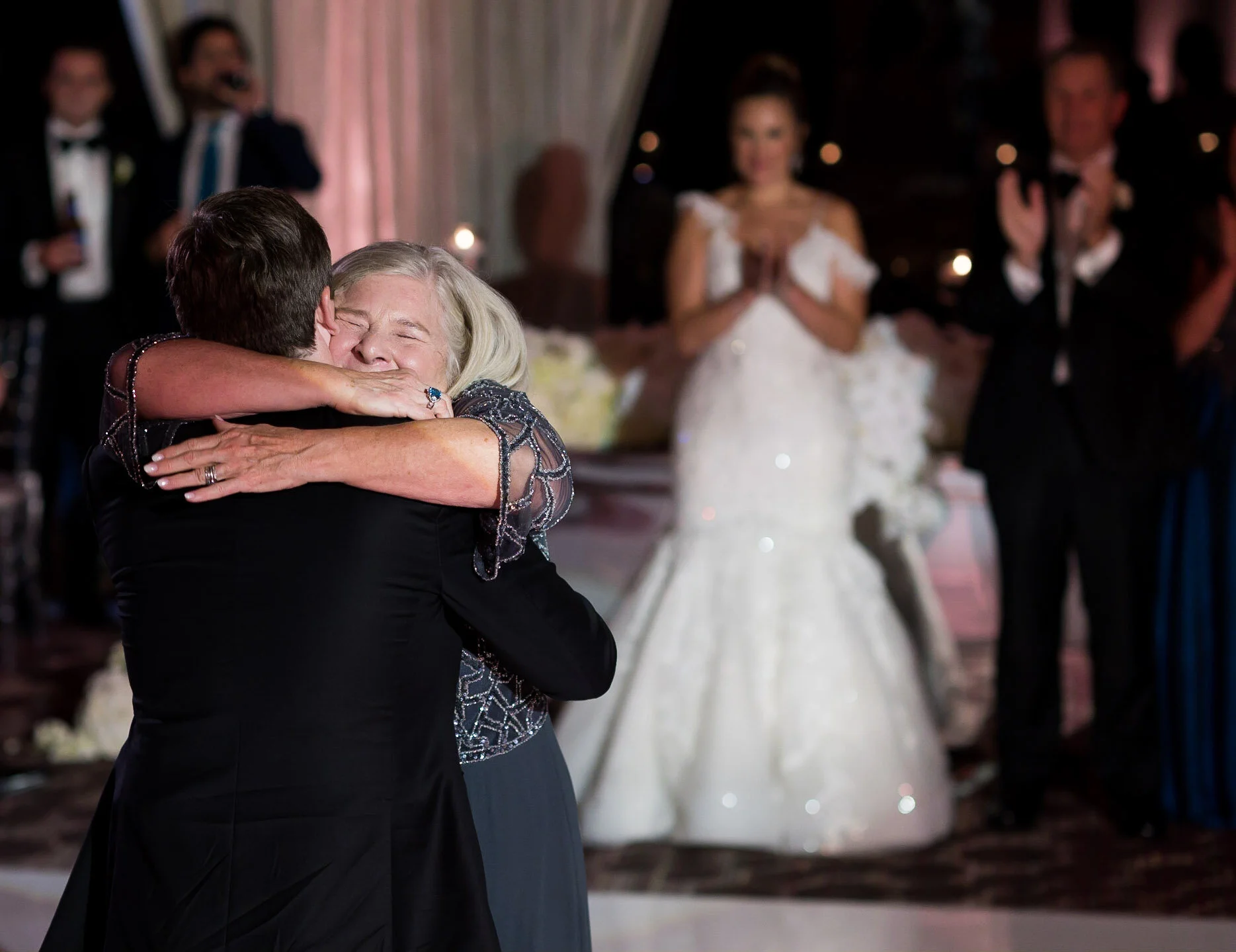 A couple is dancing closely, sharing an emotional moment at a wedding reception. The woman has grey hair and is wearing a black dress with embellishments, while the man has brown hair and is wearing a black suit. In the background, other guests, incl