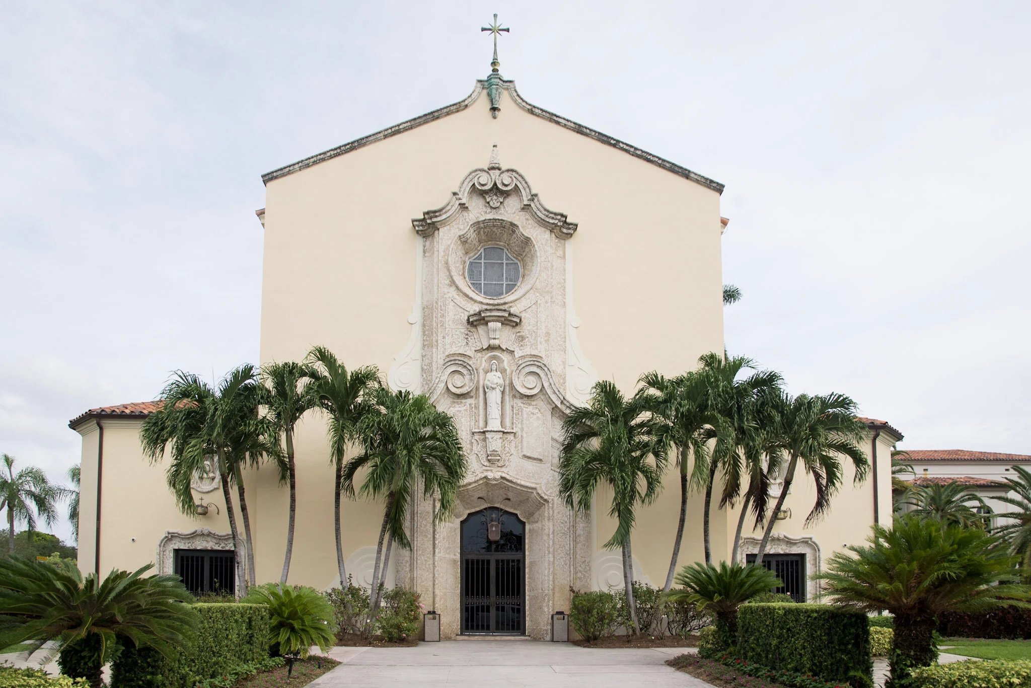 Front view of a church with cream-colored walls, ornate stone decorations, a large arched doorway, and a cross on top. Tall palm trees surround the entrance.