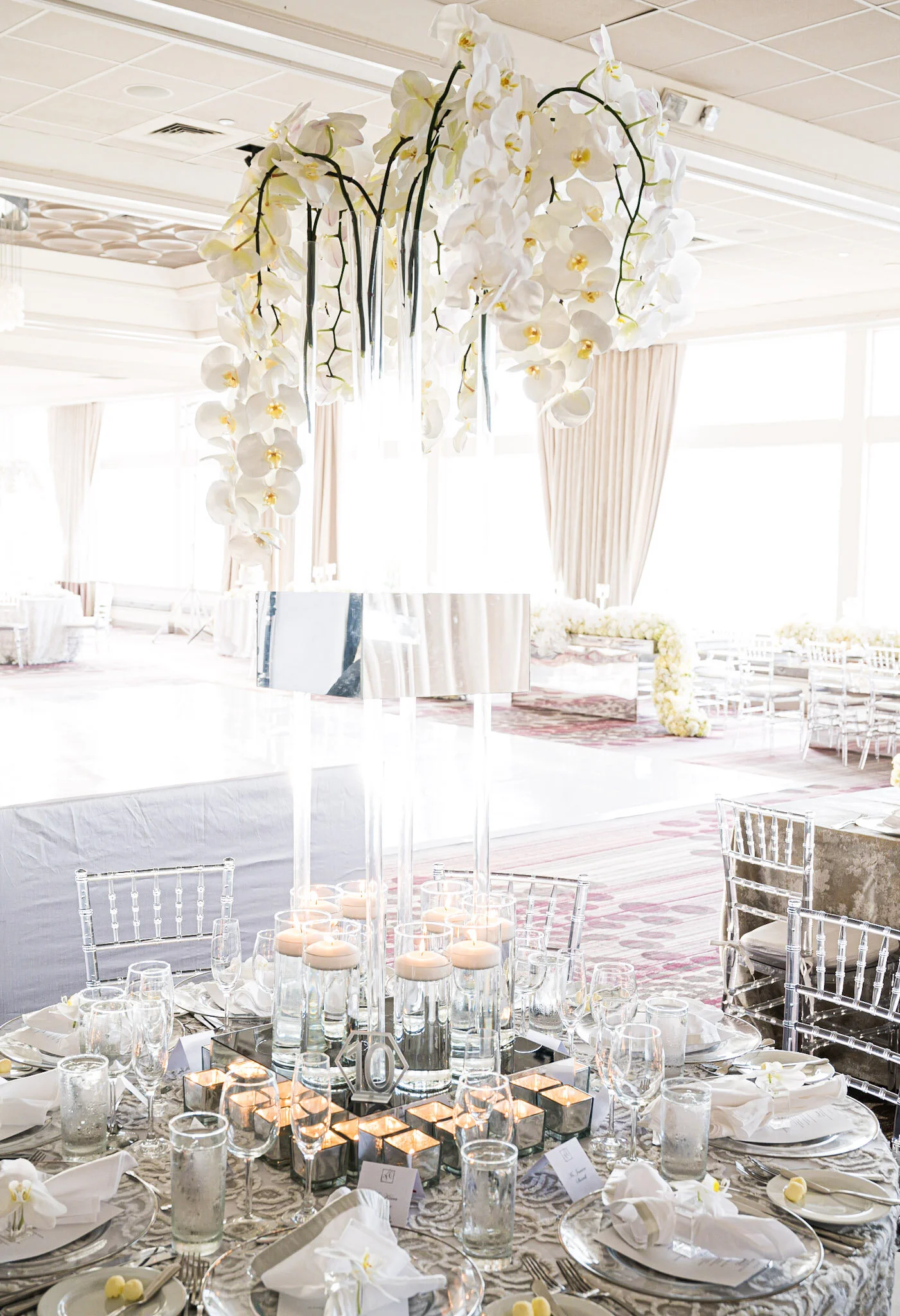 A wedding reception setup with a round table decorated with candles, glasses, and napkins, surrounded by transparent chairs, and a floral centerpiece. The background shows a bright room with large windows and curtains.
