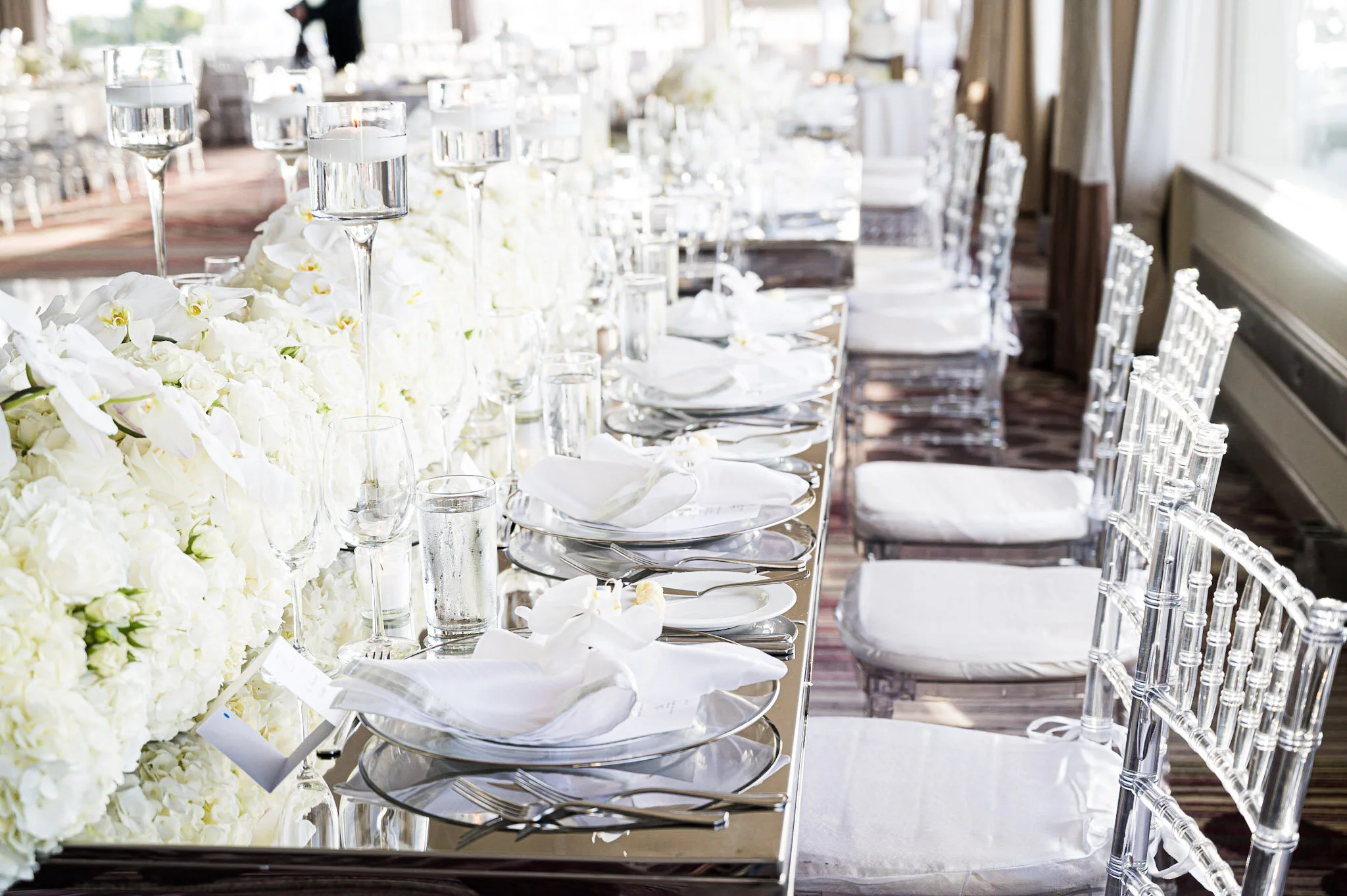 Elegant wedding reception table set with white floral arrangements, water glasses, and clear acrylic chairs in a bright room with large windows.