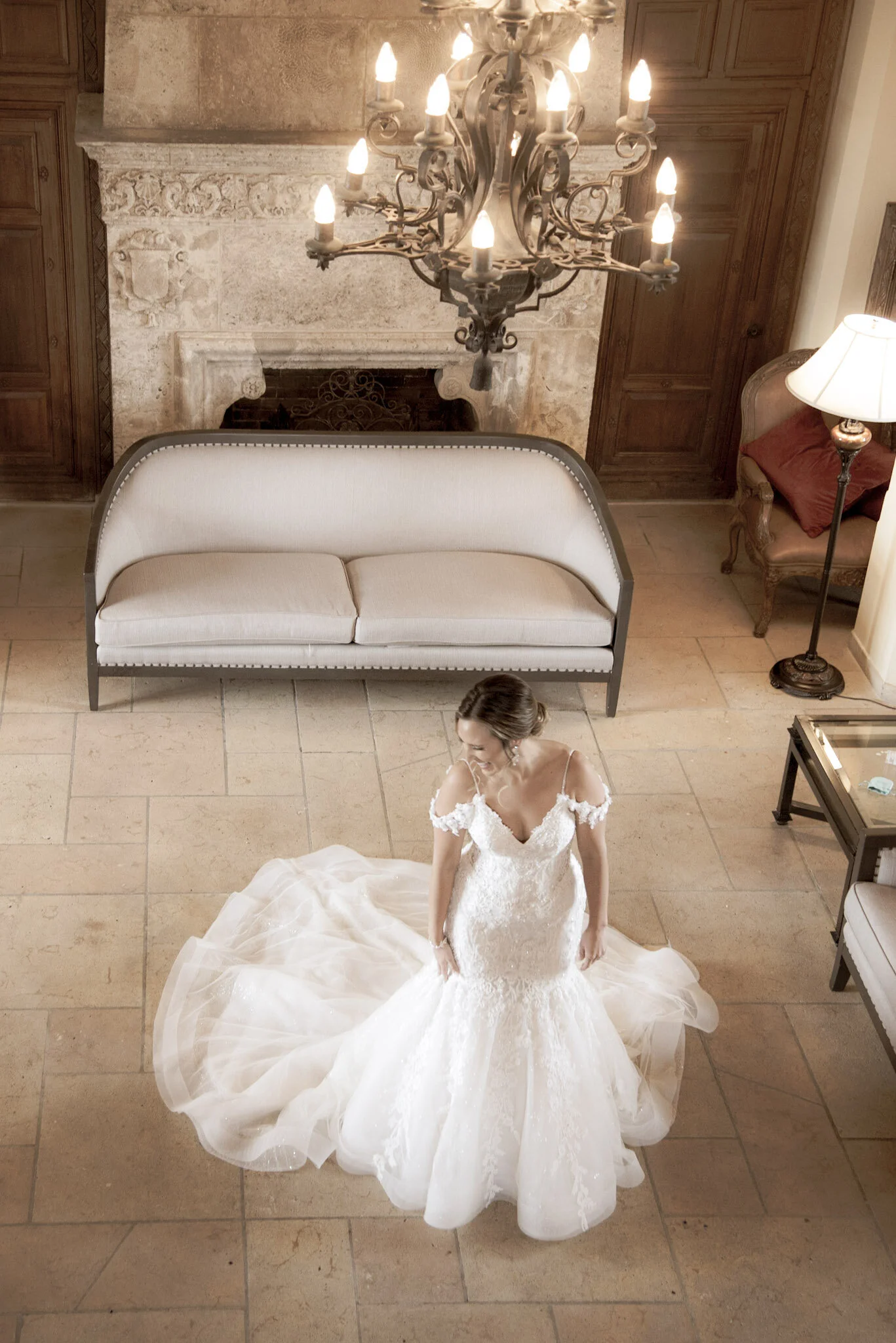 A bride standing in a room with stone flooring, wearing a white wedding dress with lace detailing and a long train, with her hair styled in an updo. The room has a fireplace with a decorative mantel, a large chandelier hanging from the ceiling, a whi