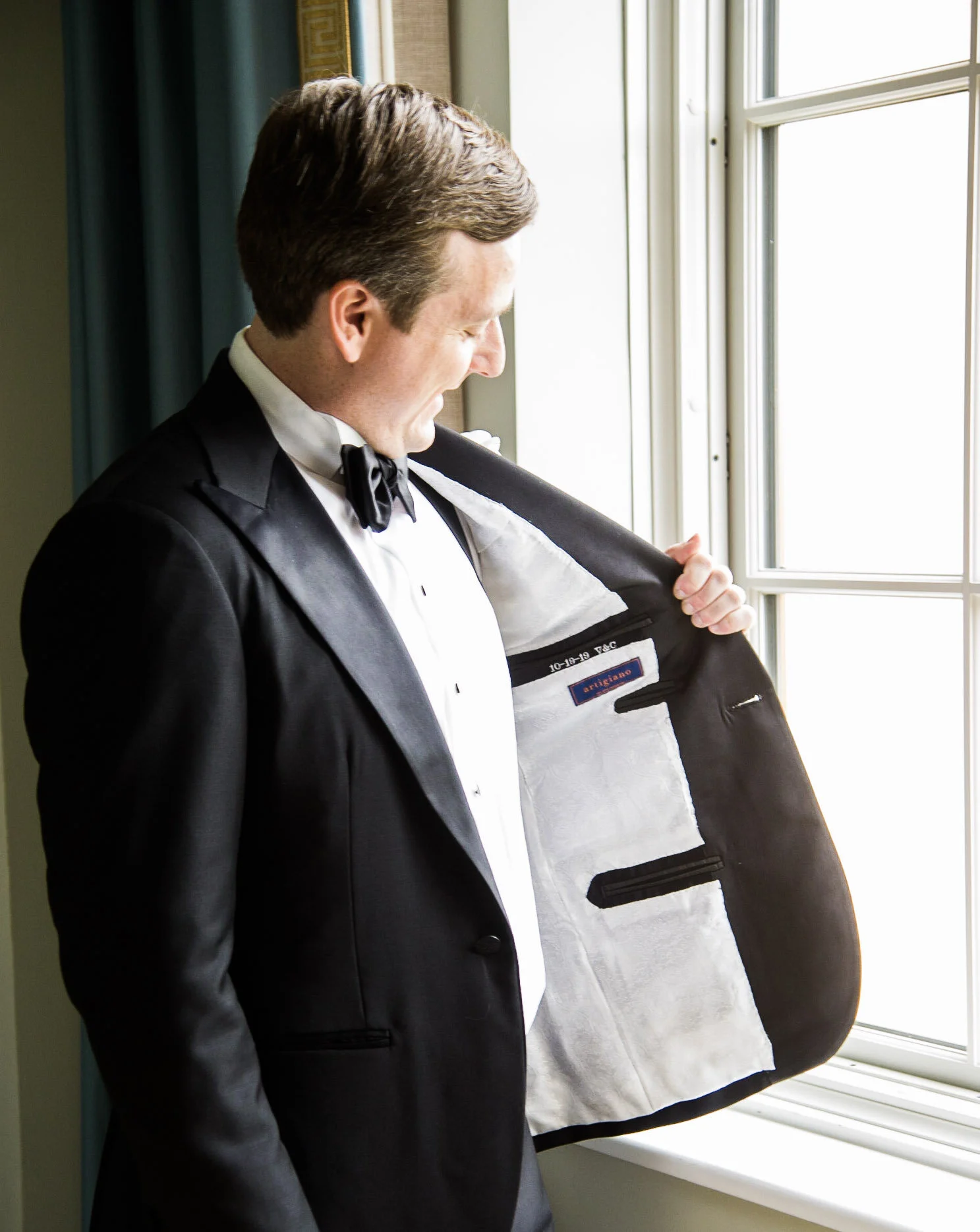 A man in a tuxedo looking out a window, holding open his jacket to reveal an interior label.