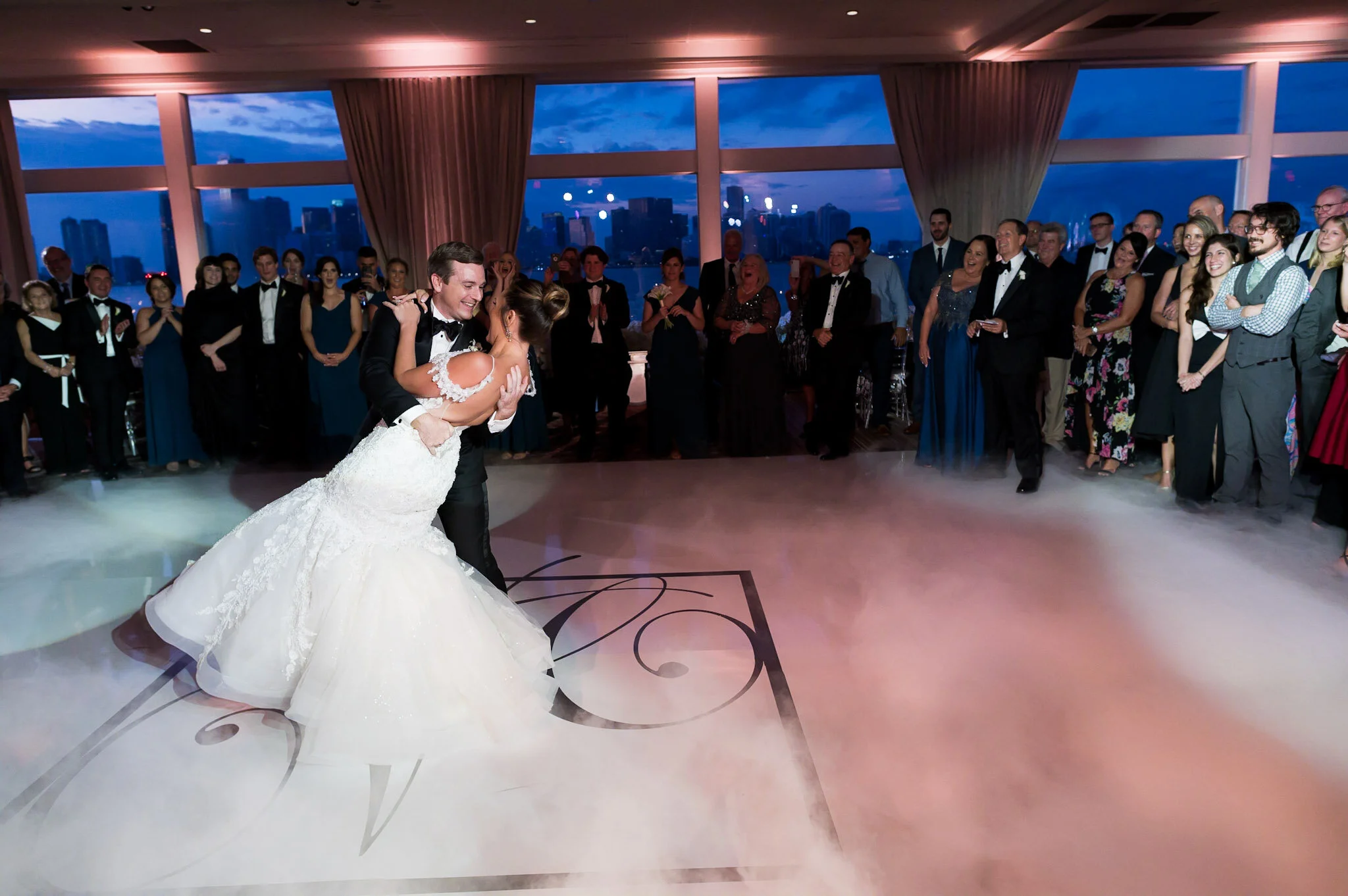 Bride and groom dancing during their wedding reception with guests watching in a spacious indoor venue with large windows showing city buildings at dusk.