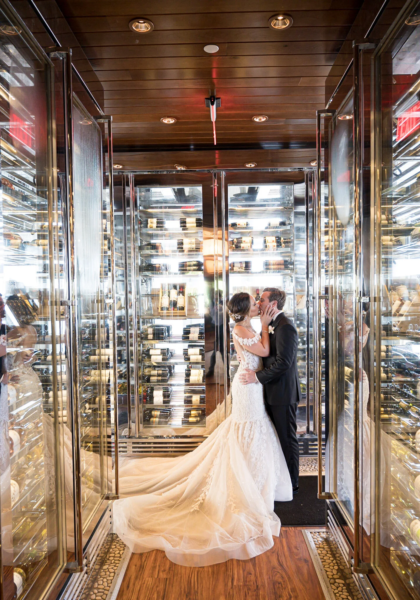 A bride and groom sharing a kiss inside a wine cellar with glass door wine refrigerators on both sides.