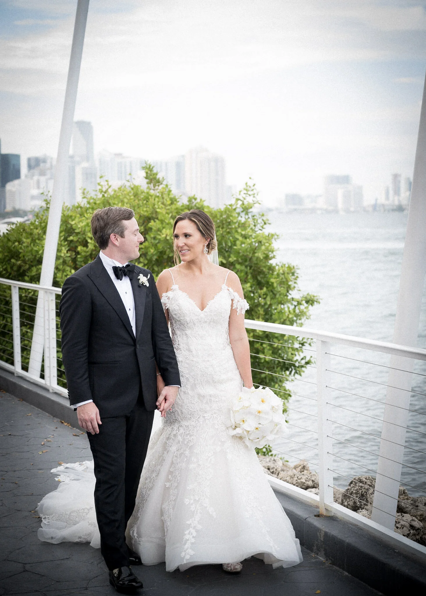A bride and groom standing outdoors near a body of water during their wedding, holding hands and looking at each other, with a city skyline in the background.
