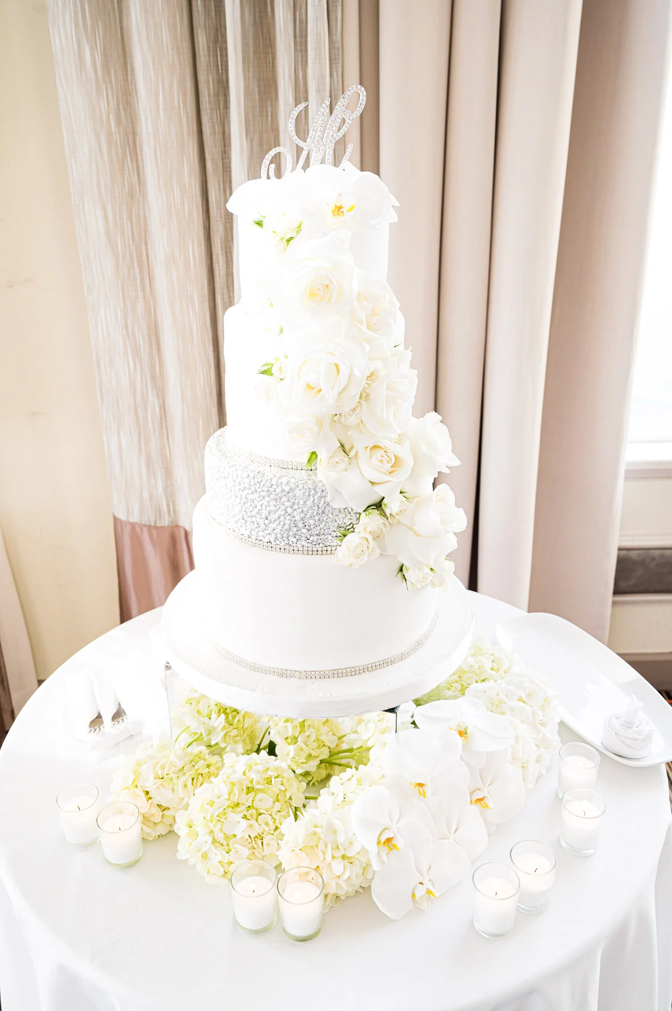 A three-tiered white wedding cake decorated with white roses and orchids, topped with a silver cake topper that reads 'Love'. The cake sits on a table surrounded by white flowers and candle votives.