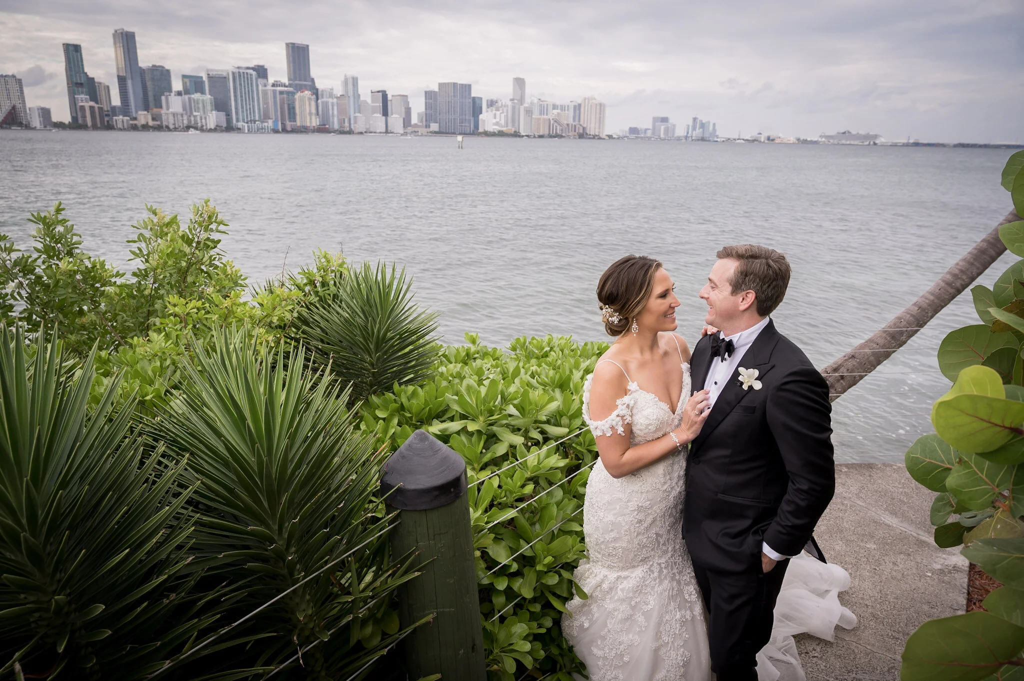 A bride and groom smiling and gazing at each other outdoors near water with a city skyline in the background, surrounded by green plants and trees.