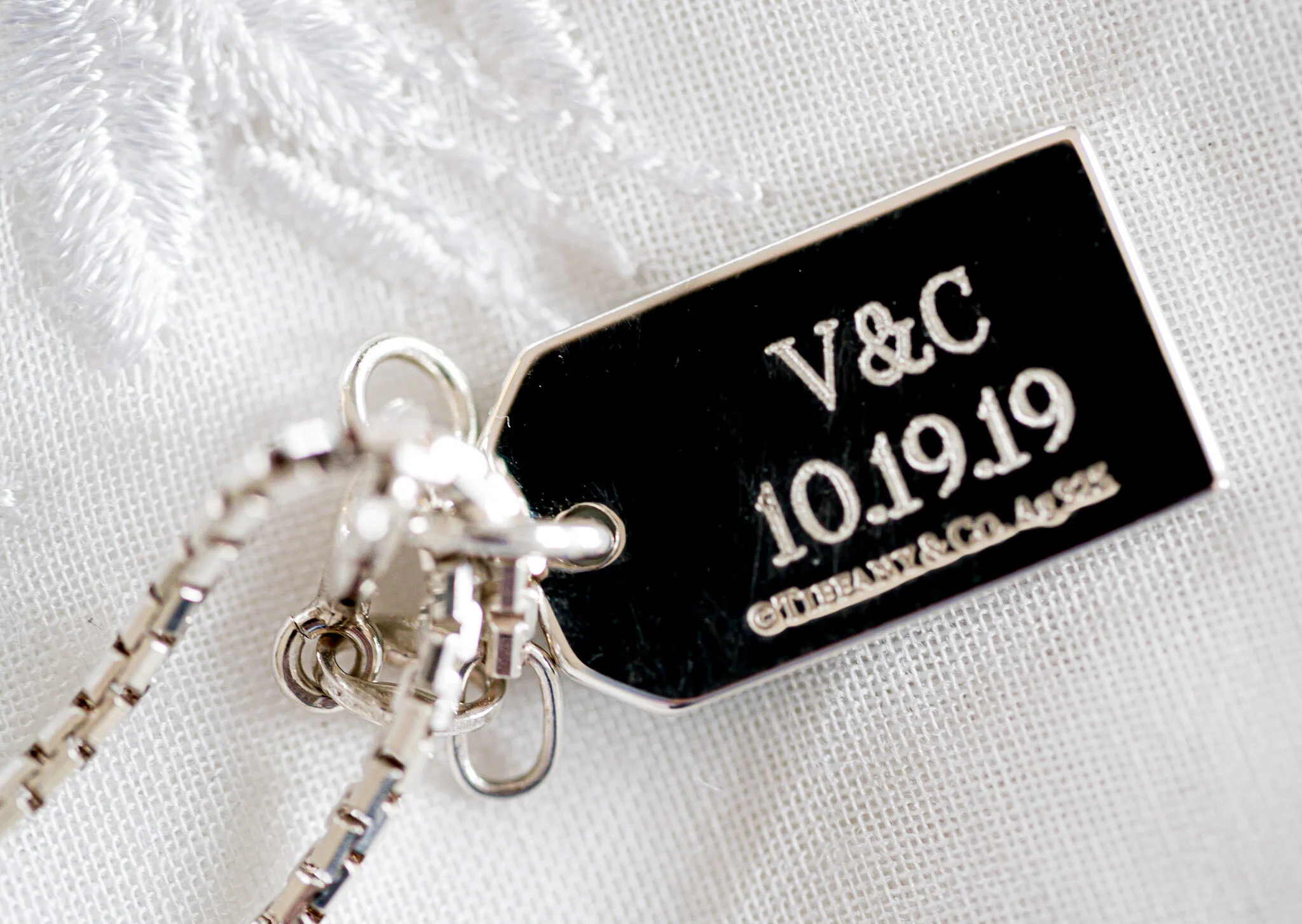 A close-up of a silver keychain with a black and silver tag that has engraved text, placed on a white textured fabric.